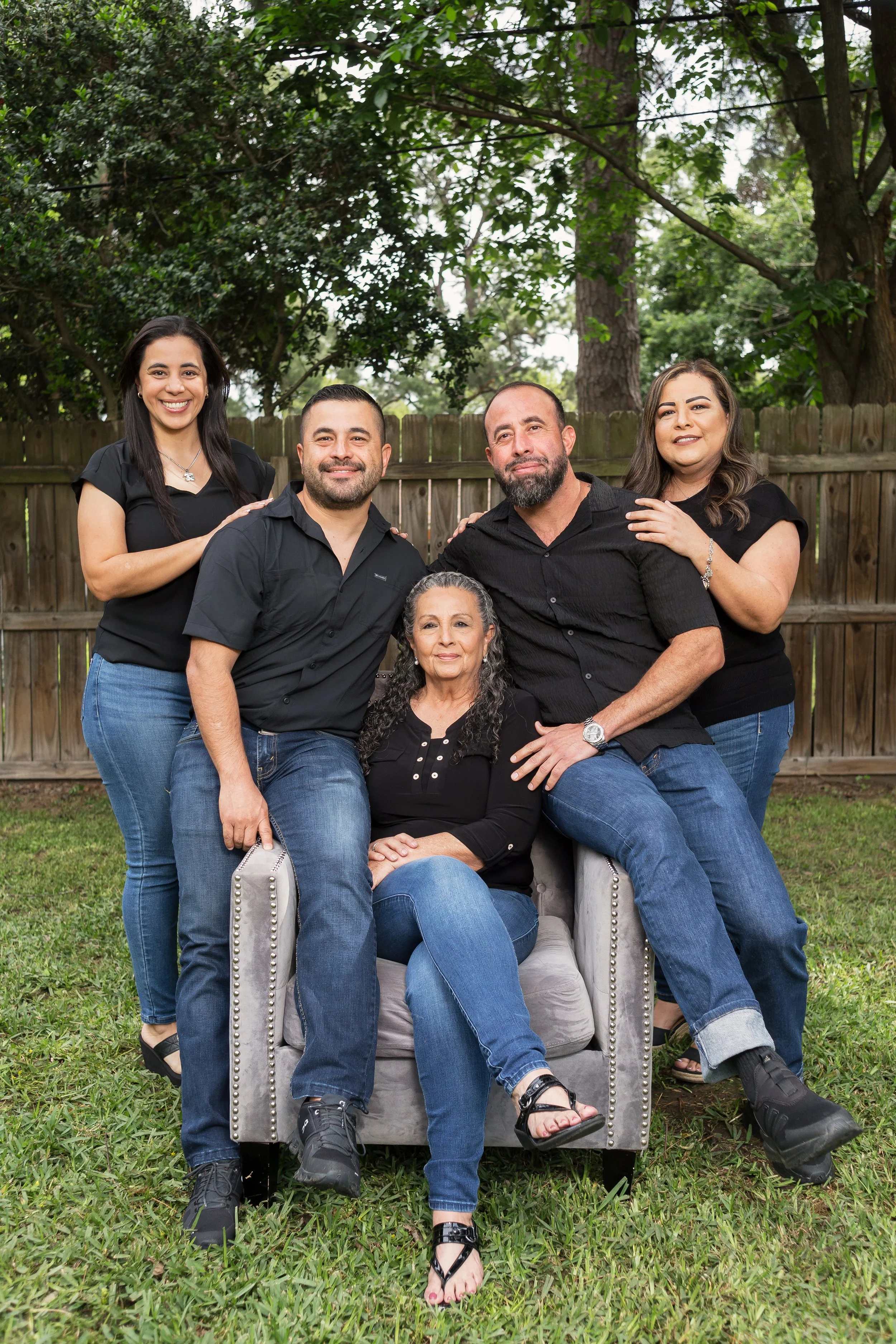 Family gathering in backyard with five adults, three women and two men, sitting and standing around a woman seated on a gray armchair, all wearing casual black and blue clothing, with a wooden fence and green trees in the background.