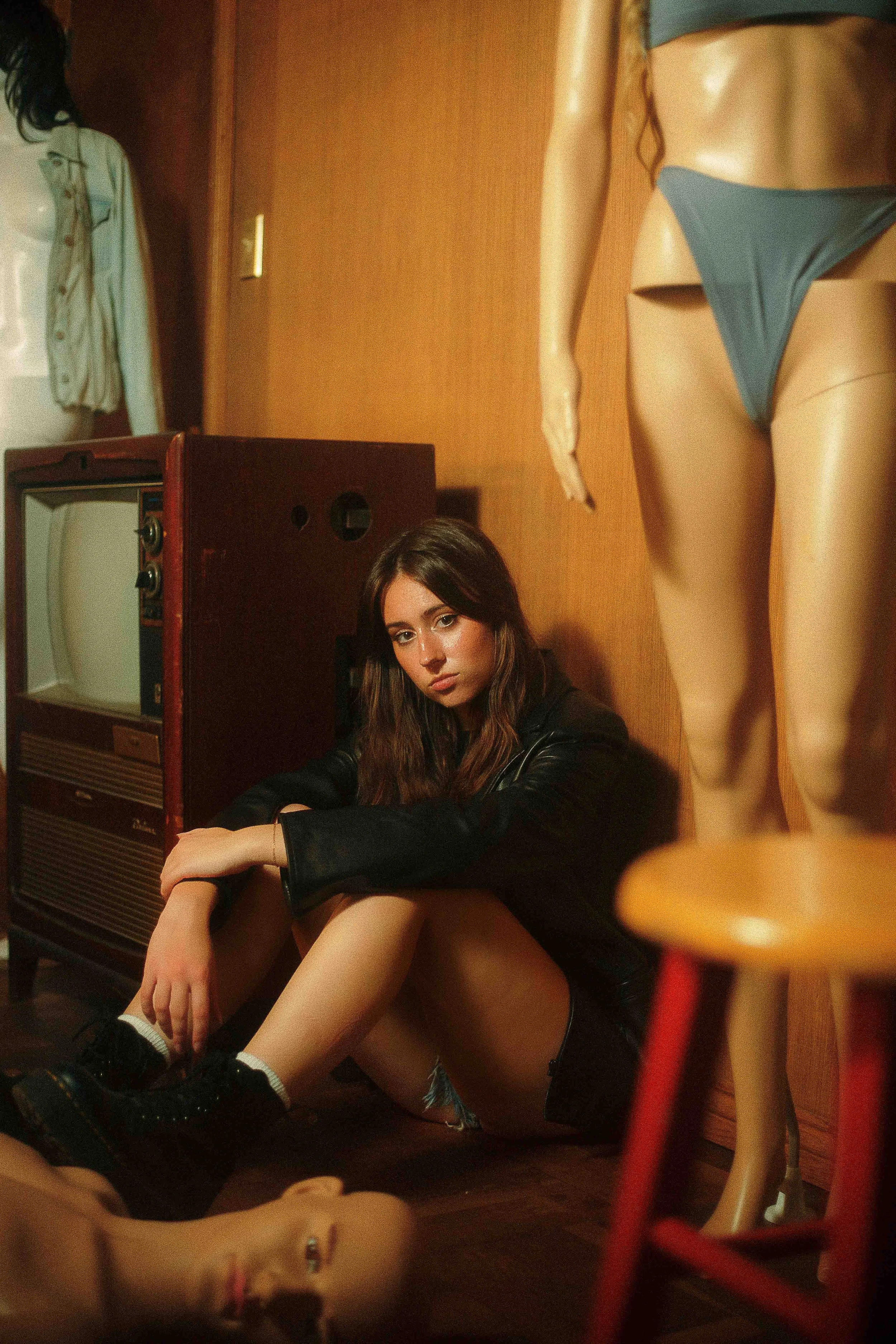 Moody indoor portrait of a young woman with brown hair sitting on the floor in a dimly lit room, wearing a black jacket, denim shorts, and black boots, surrounded by vintage objects including a mannequin in a blue bikini, a retro television, and a wo