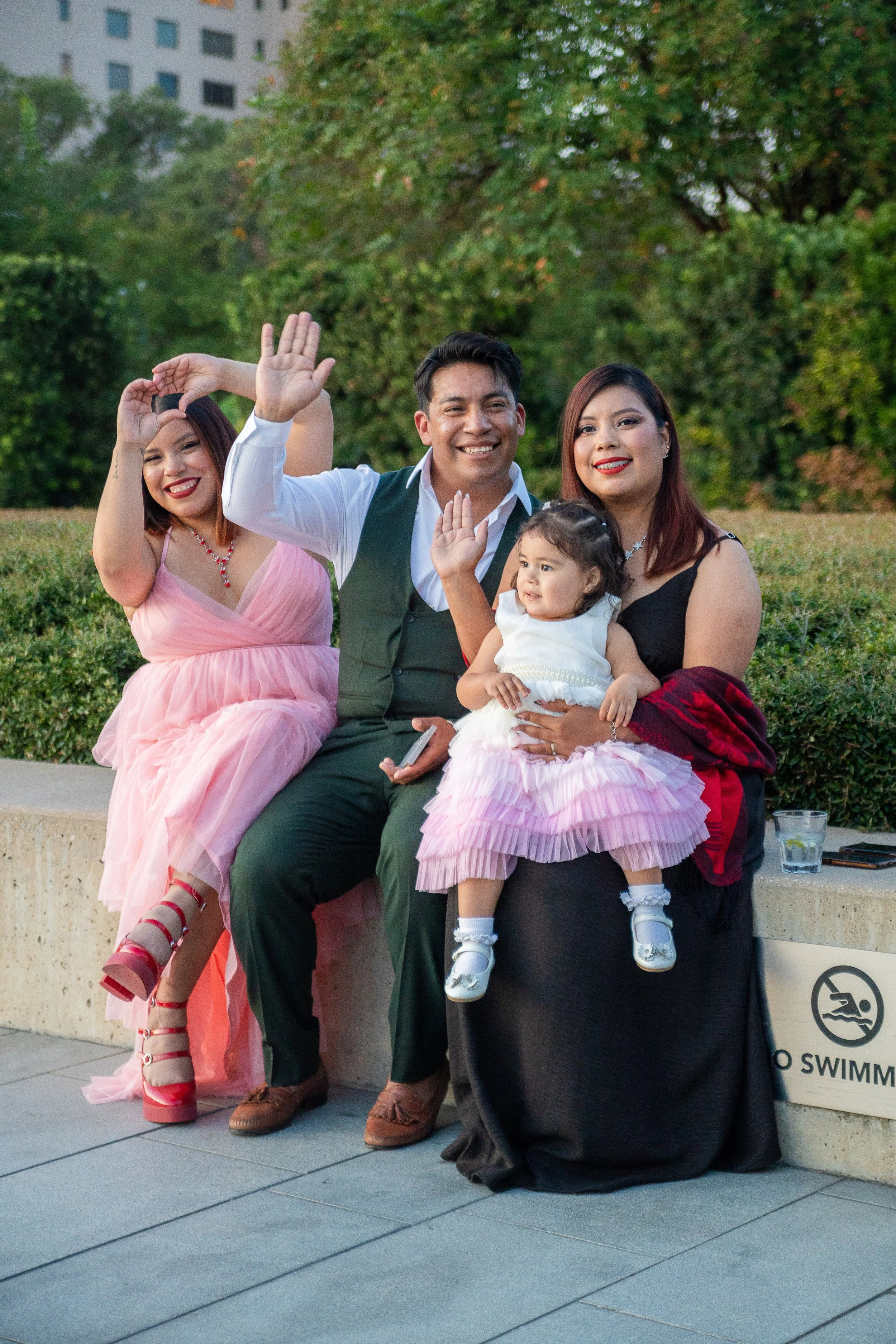 A group of four people, including three women, a man, and a young girl, sit on a concrete bench outdoors, smiling and waving at the camera. They are in a park or garden area with trees and a building in the background. The woman on the left is in a p