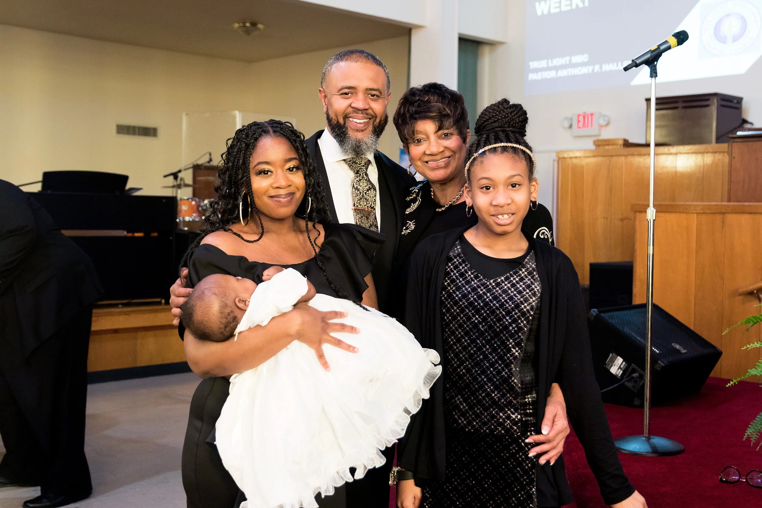 Family gathered in church, with a woman holding a baby, a man and woman standing close, and a girl in front. They are smiling and dressed in formal attire, with a stage and musical equipment in the background.