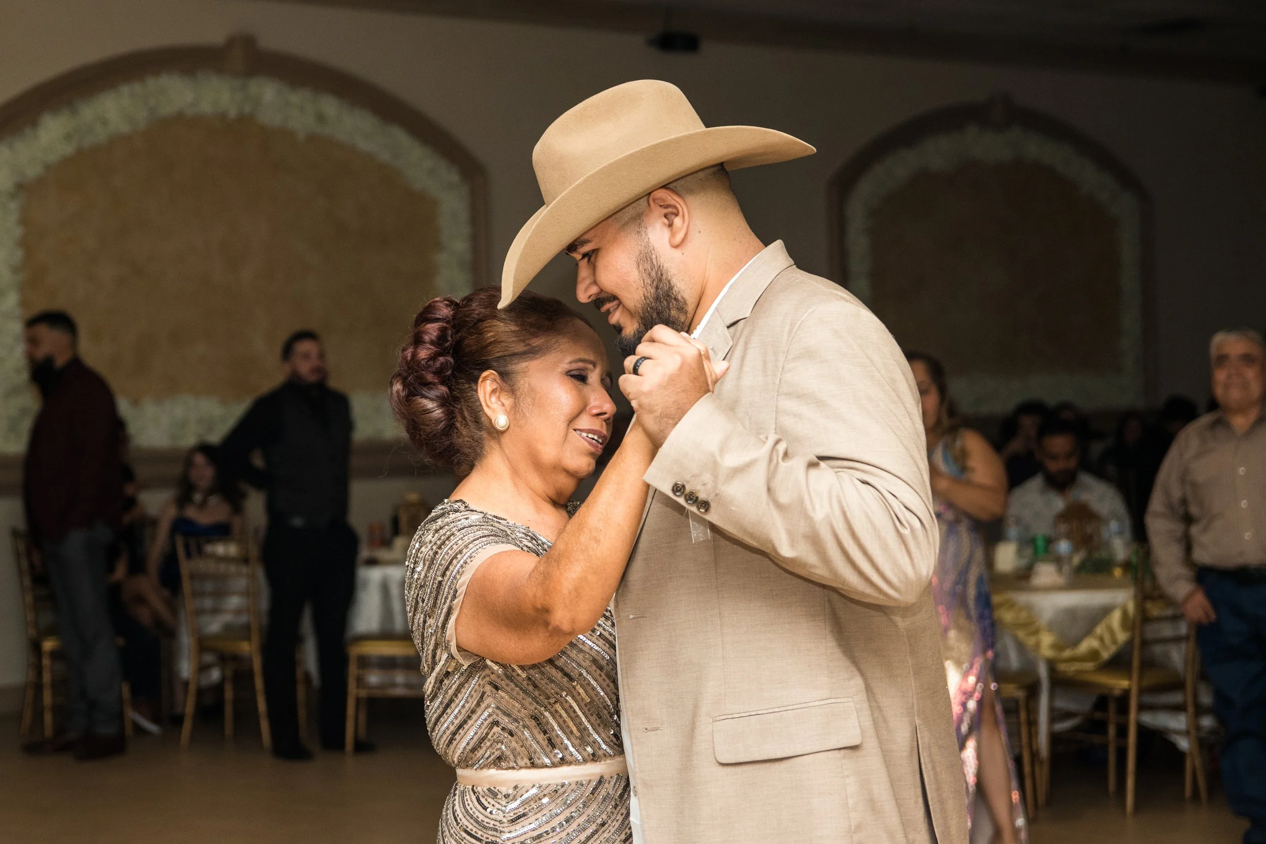 A man and woman dancing closely at a celebration, with the man wearing a light suit and wide-brimmed hat, and the woman in a patterned dress, both smiling and embracing.
