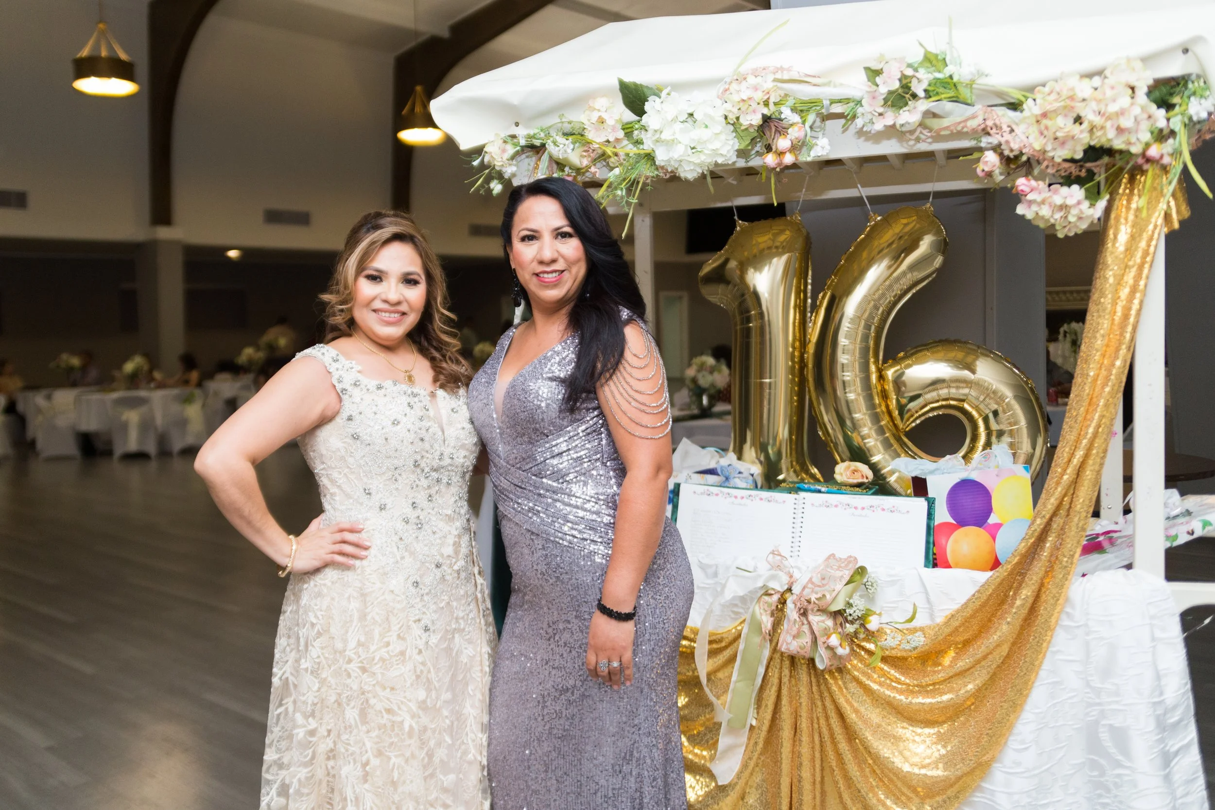 Two women dressed in formal attire posing at a celebration, with large gold balloons spelling out 16, and decorated table with flowers and balloons in the background.