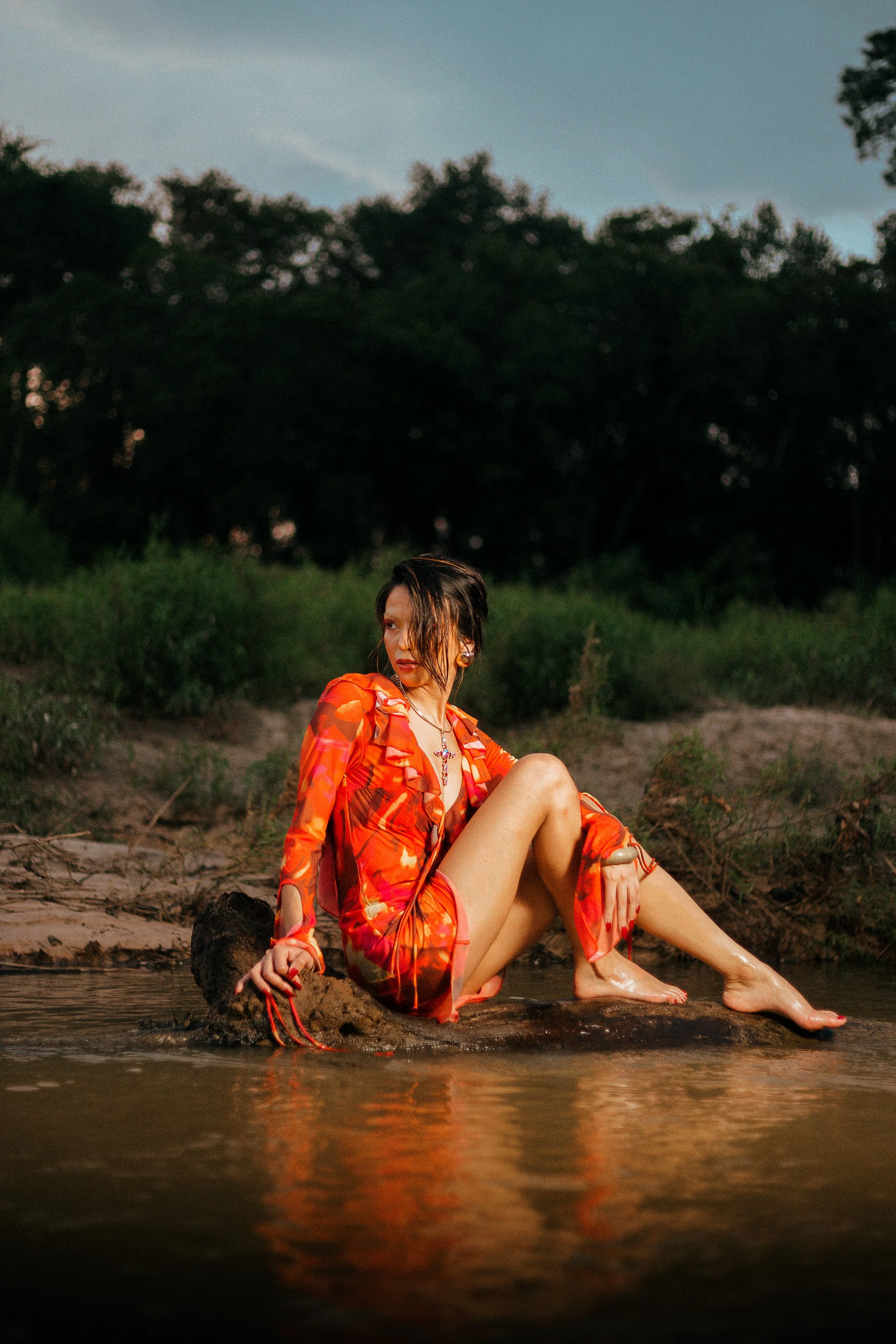 A woman in an orange and red floral dress sitting on a rock in a stream, with dark green trees in the background during dusk.