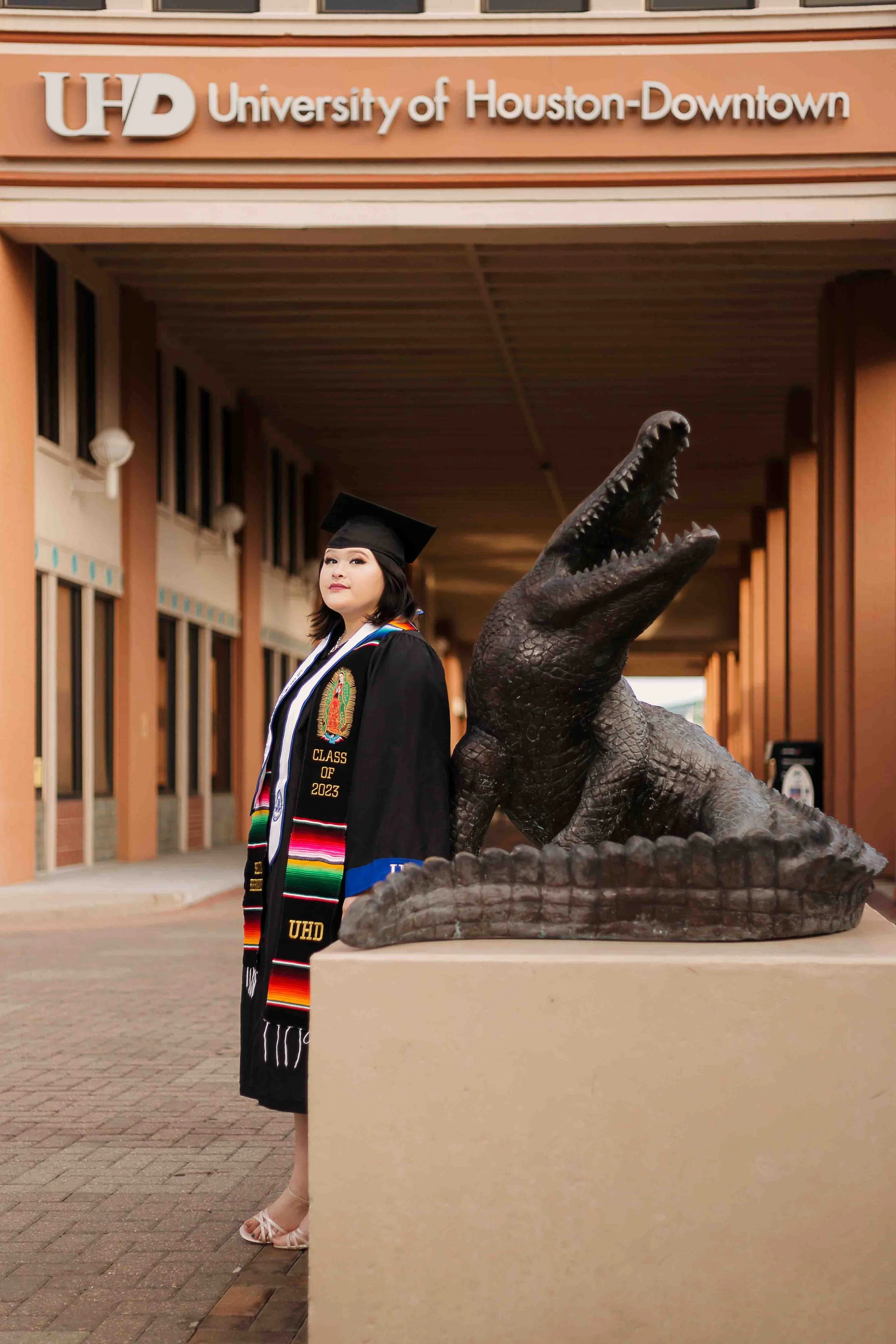 University of Houston–Downtown graduation portrait featuring a graduate in cap and gown standing outdoors near a statue of a crocodile with its mouth open on campus