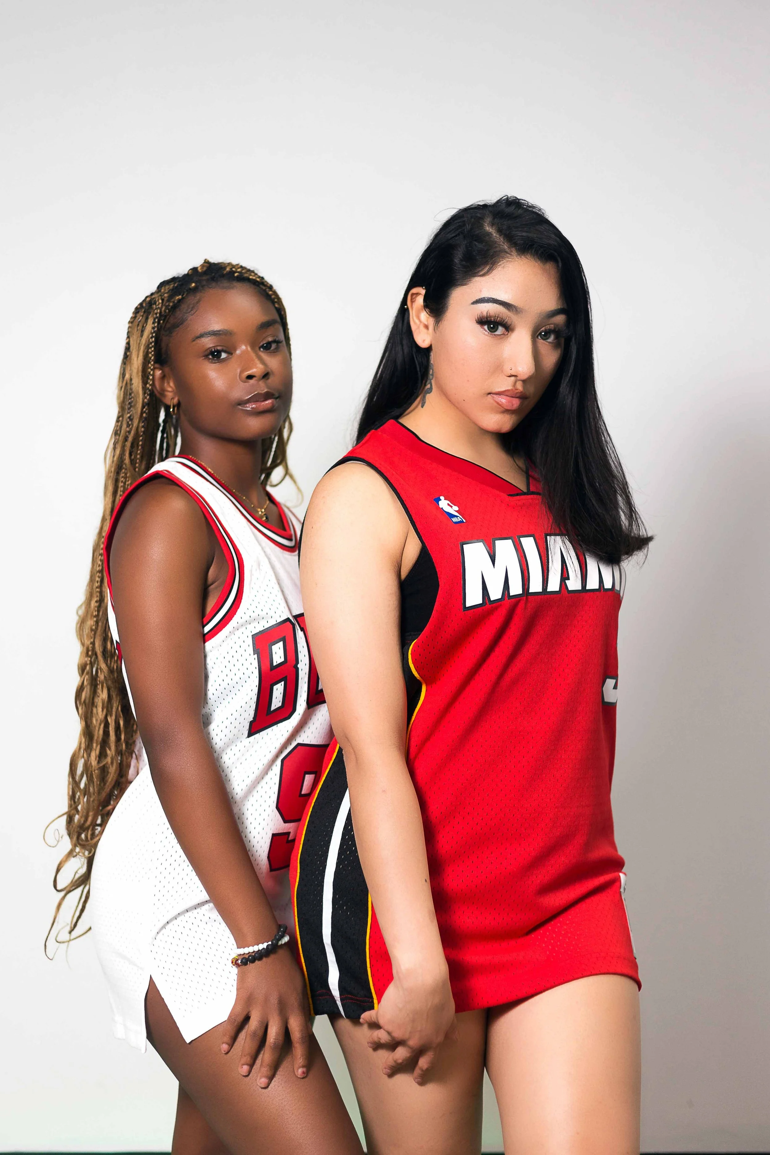 Retail store portrait featuring two women wearing Miami Heat basketball jerseys — one in white and one in red — standing against a clean, light-colored studio background