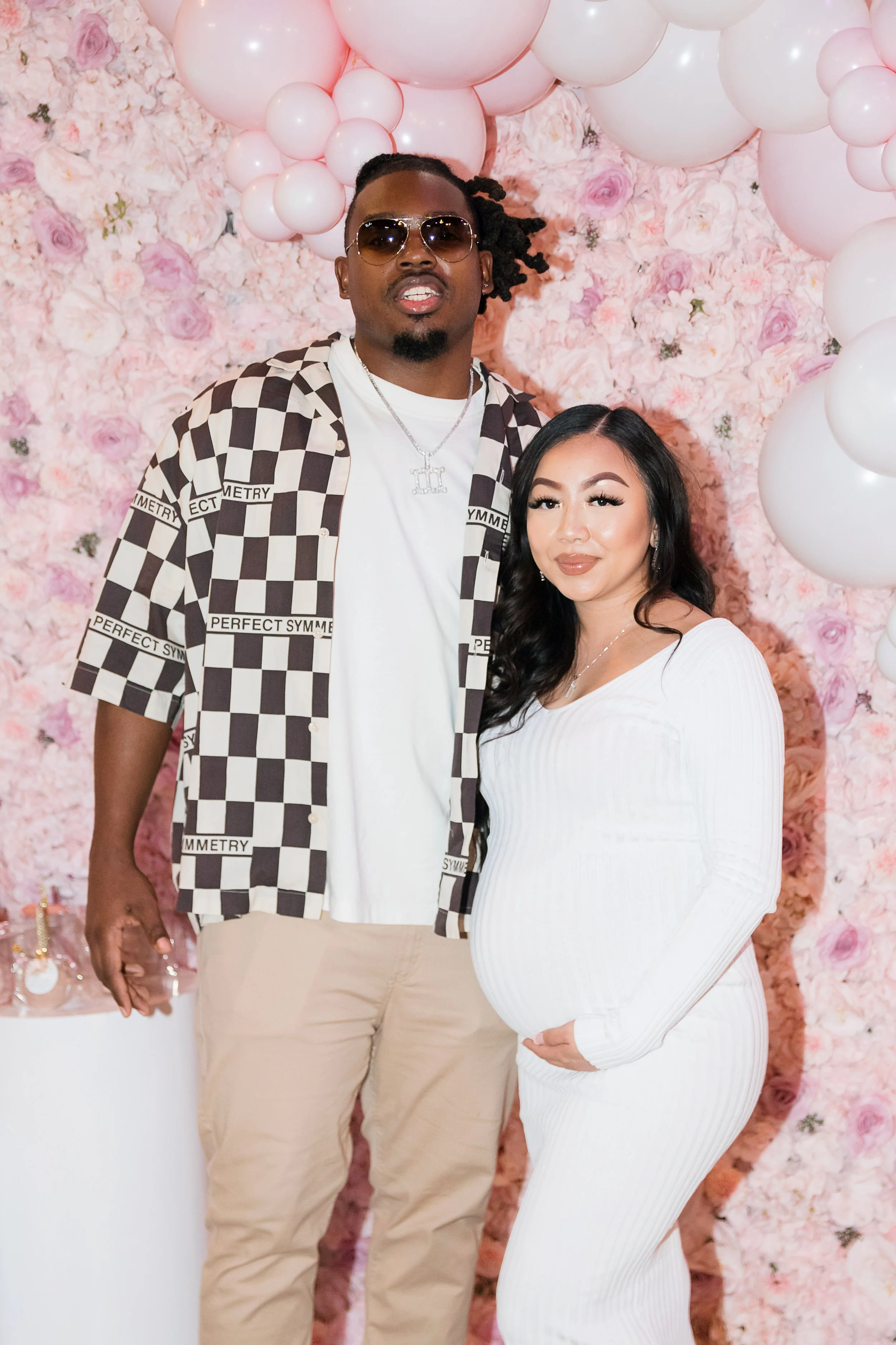 A man and a pregnant woman standing together at a celebration with pink floral background and balloon decorations.
