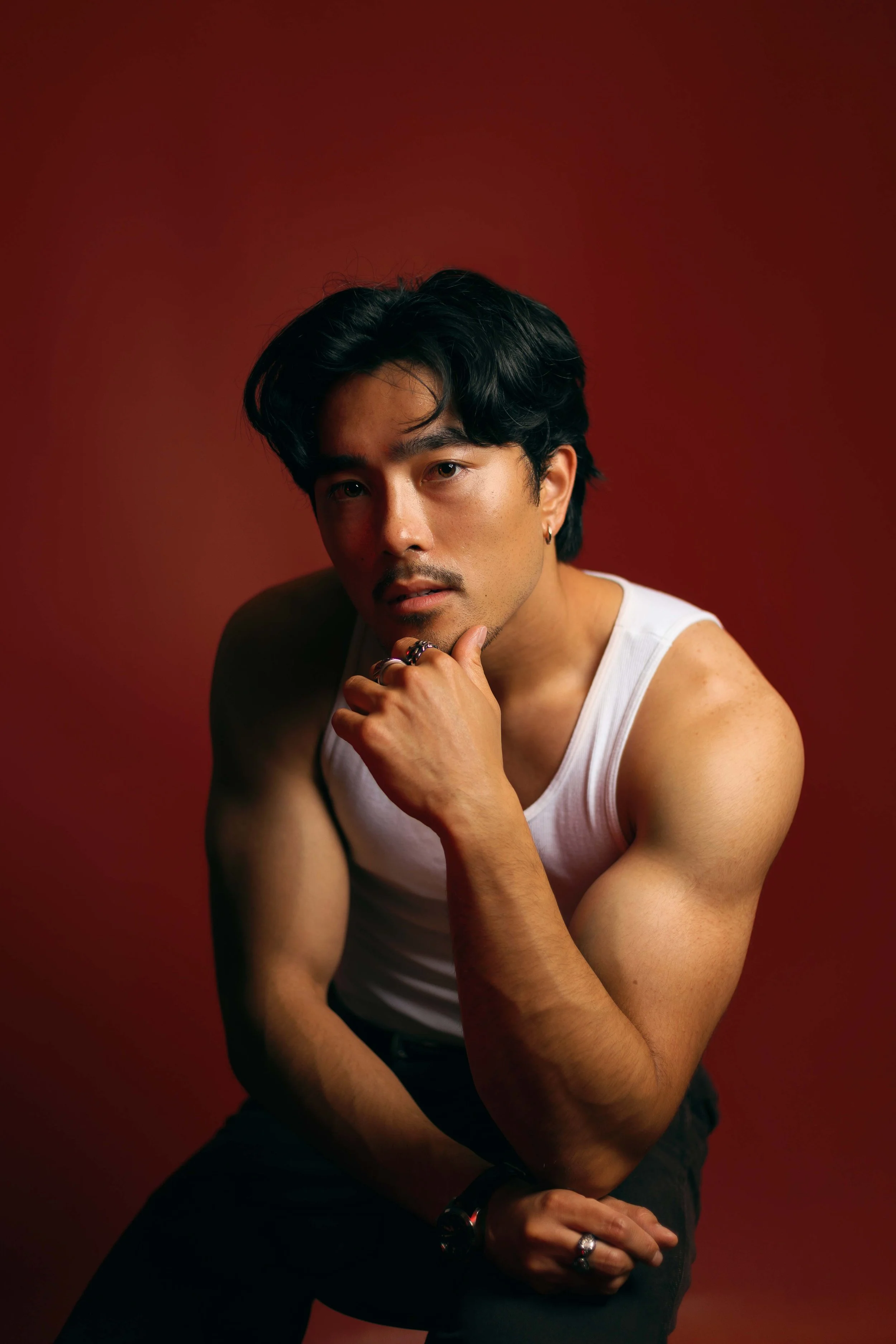 Creative editorial portrait of young man in white tank top posed against deep red background, resting chin on hand with serious expression.