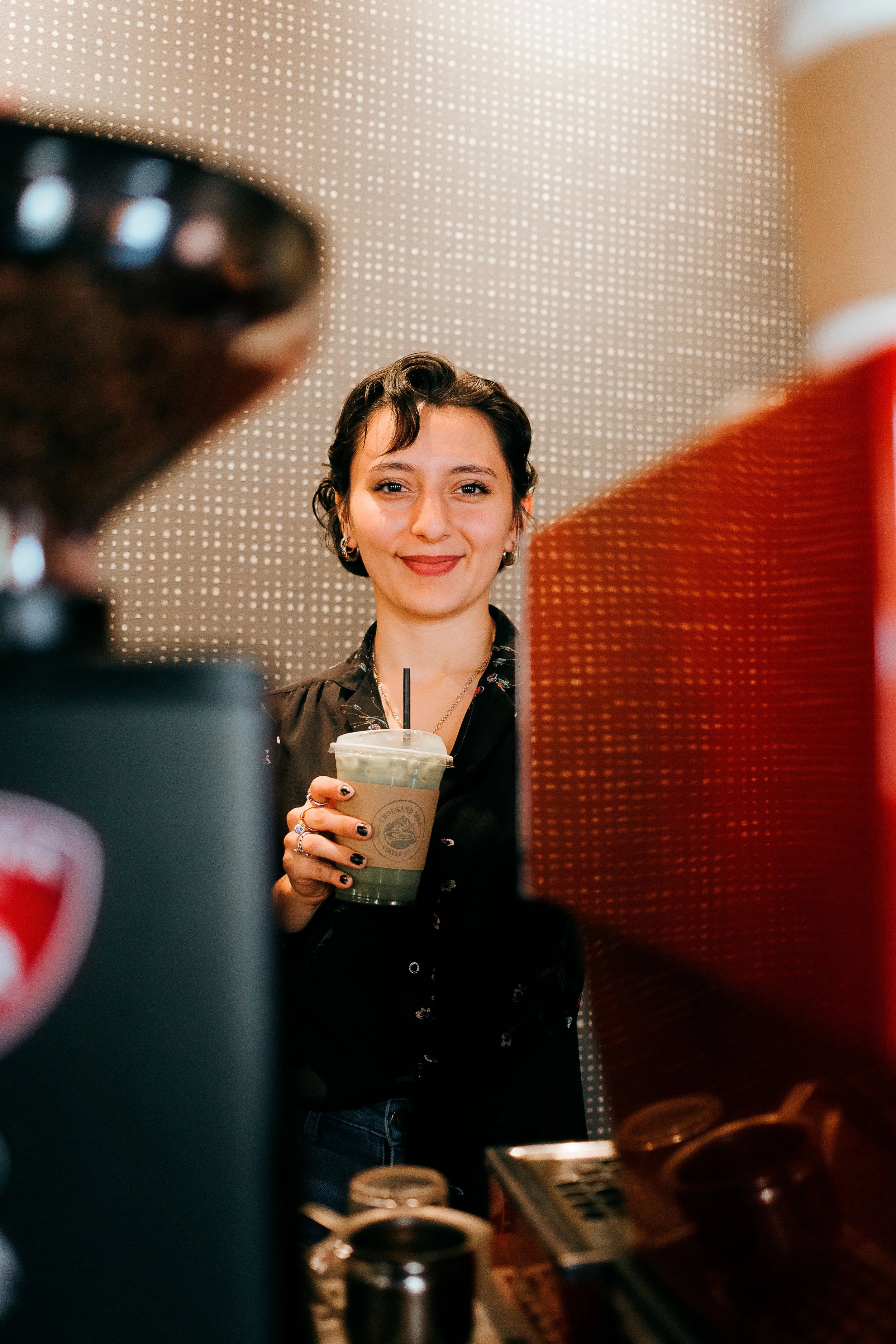 A woman with short dark hair smiling and holding a cold beverage at a coffee shop or café, seen through coffee machines and cups.