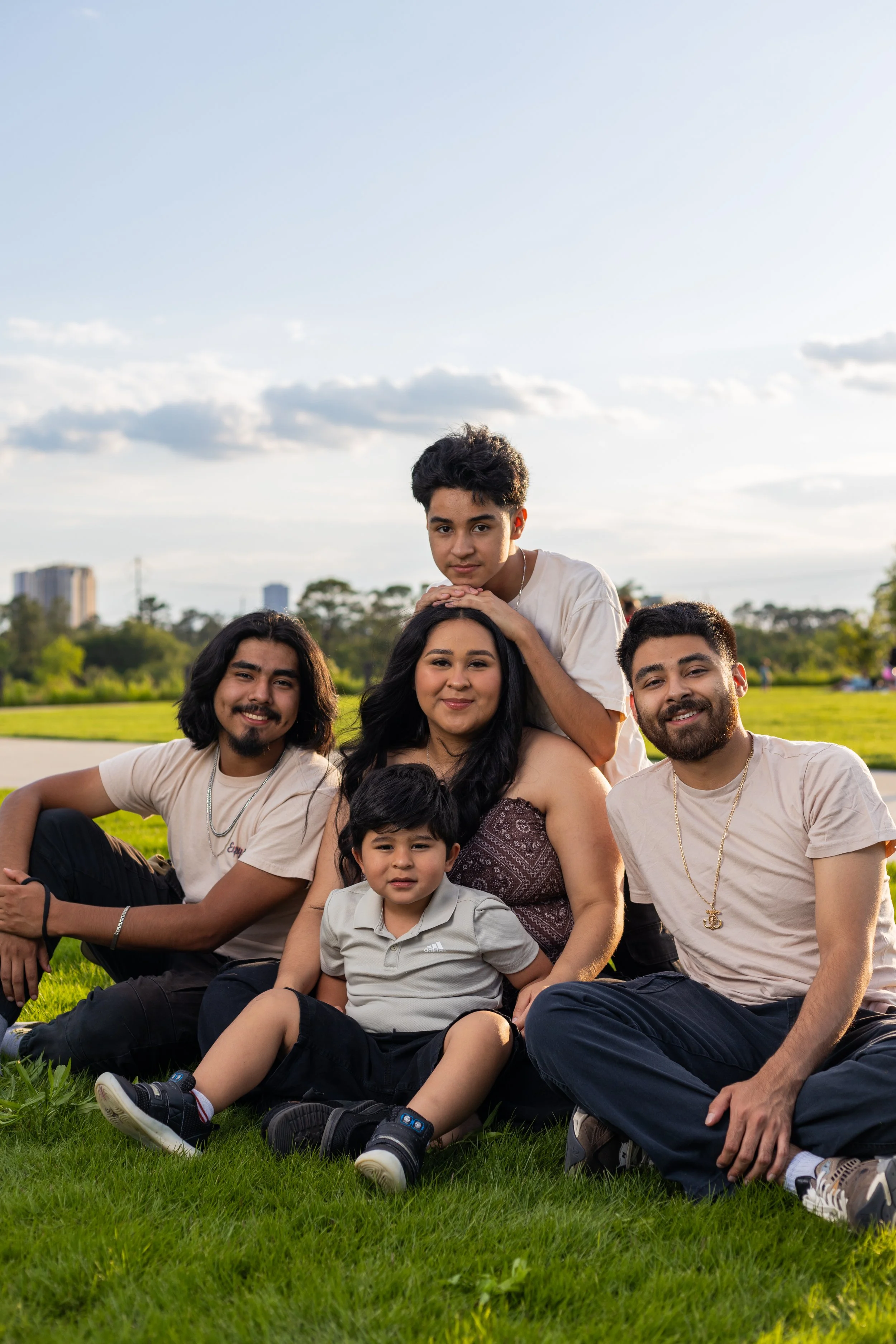 A group of six people, including children and adults, sitting on grass in a park during daytime, smiling at the camera with trees and buildings in the background.