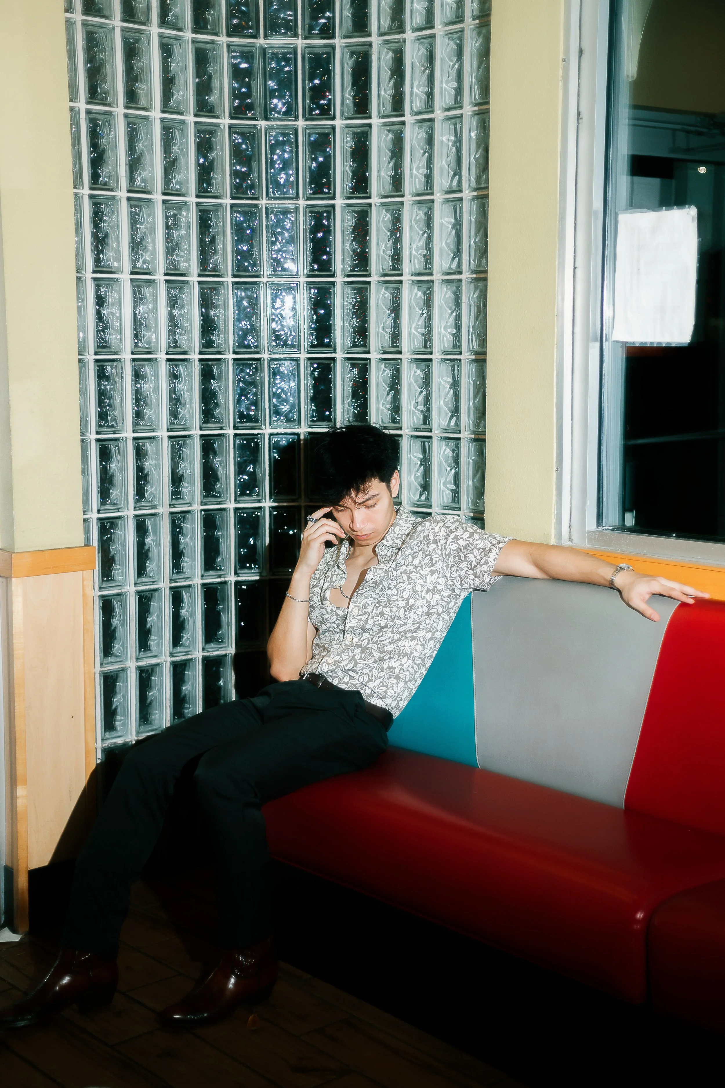 Young man sitting on a colorful postmodern bench, looking at his phone, in front of a glass block wall, wearing a patterned shirt and black pants.