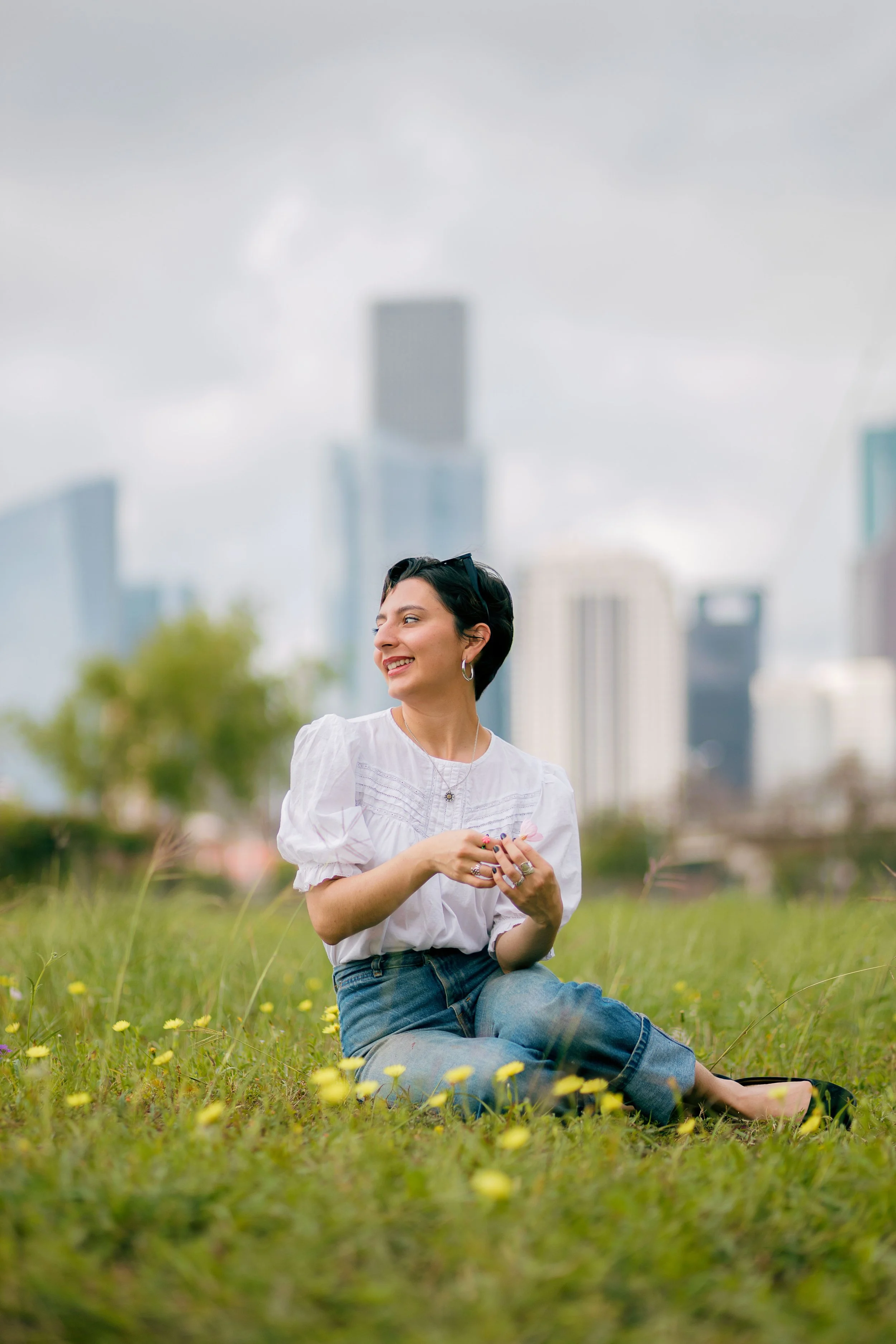 A woman sitting on grass in an urban park, smiling and looking to the side, with city skyscrapers in the background.
