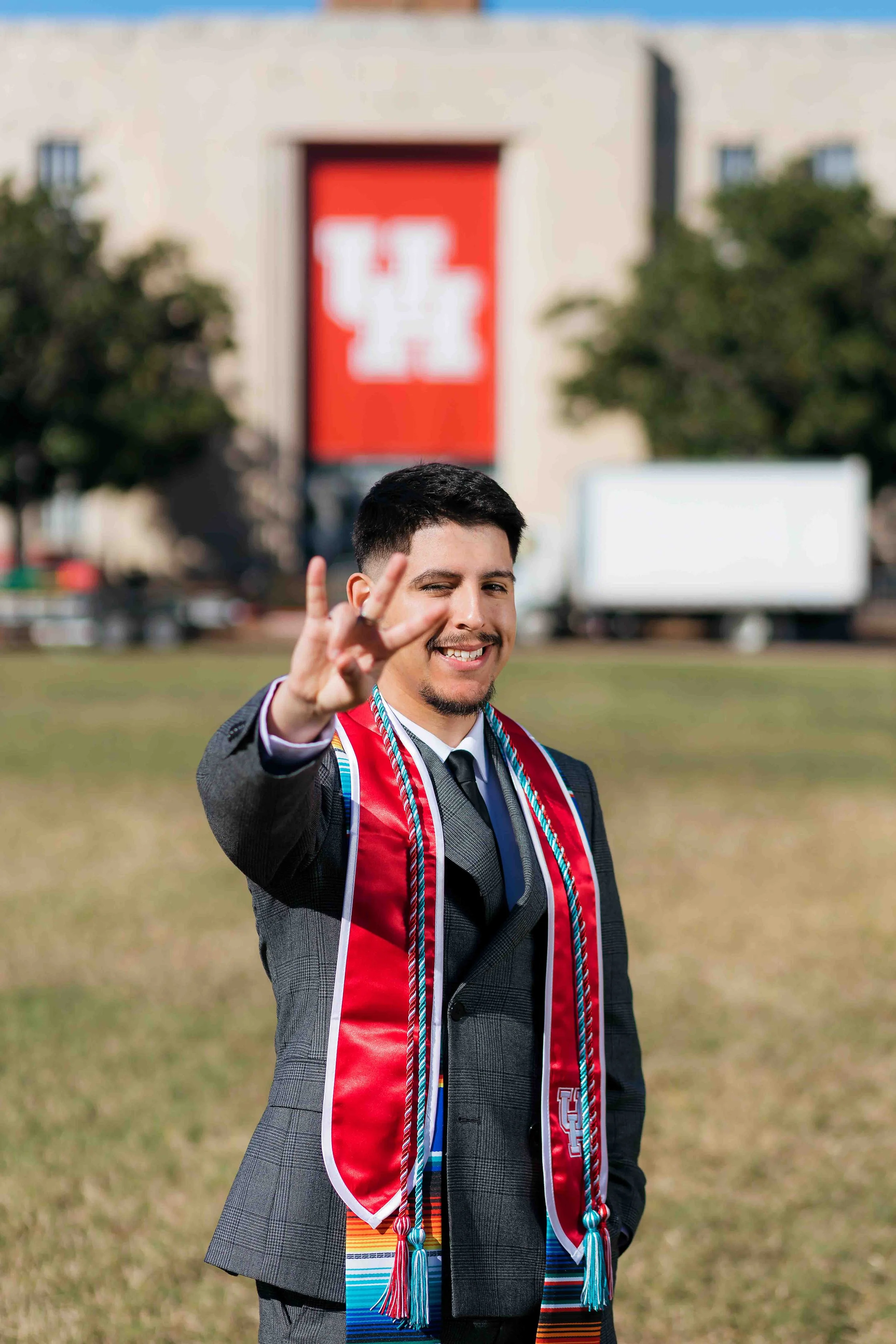 University of Houston graduate portrait featuring a young man in a gray suit making a peace sign while standing on a lawn, with trees, a university building, and a large red banner displaying the UH logo in the background