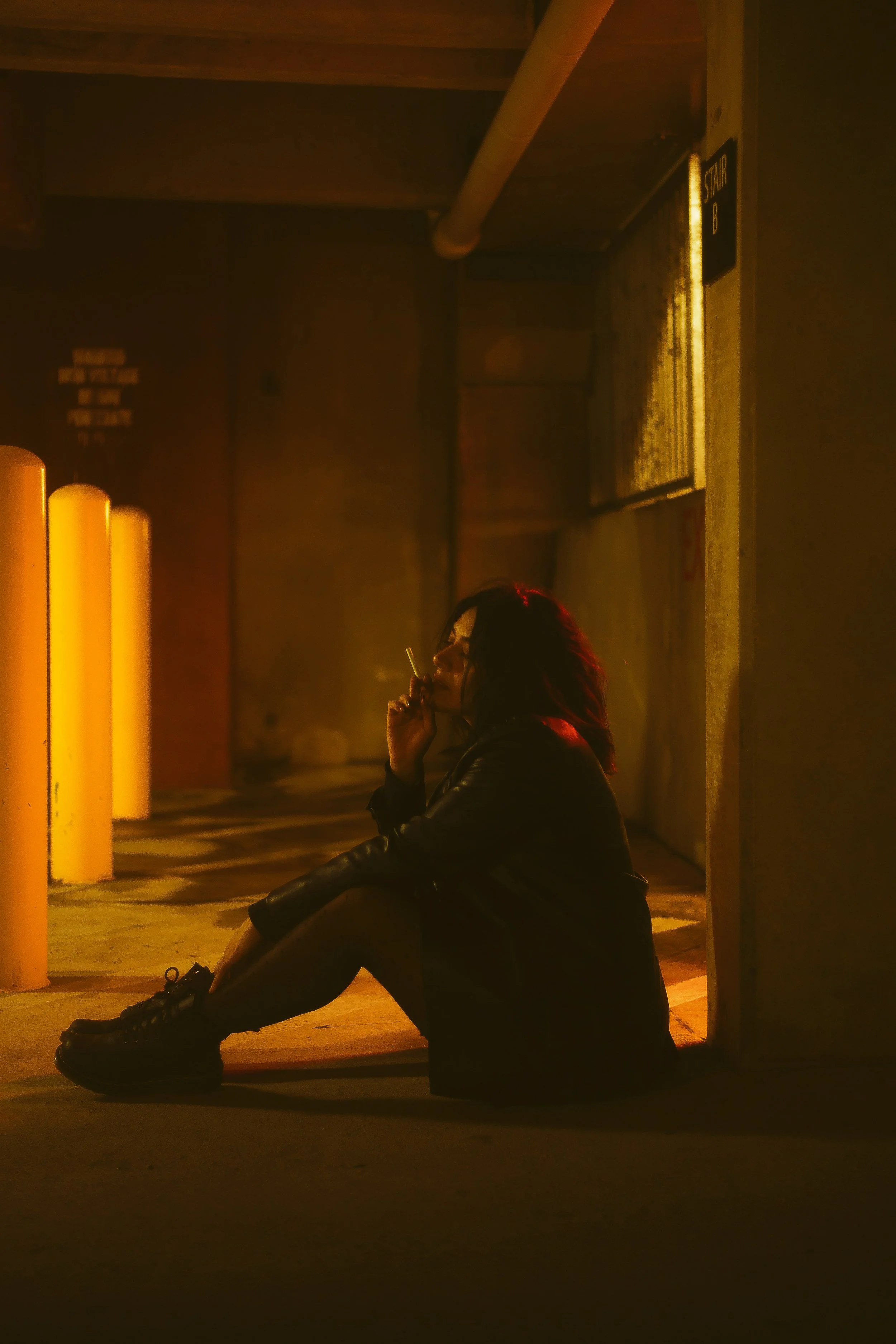 A woman sitting on the ground in an industrial parking garage at night, smoking a cigarette, illuminated by yellow-orange lighting.