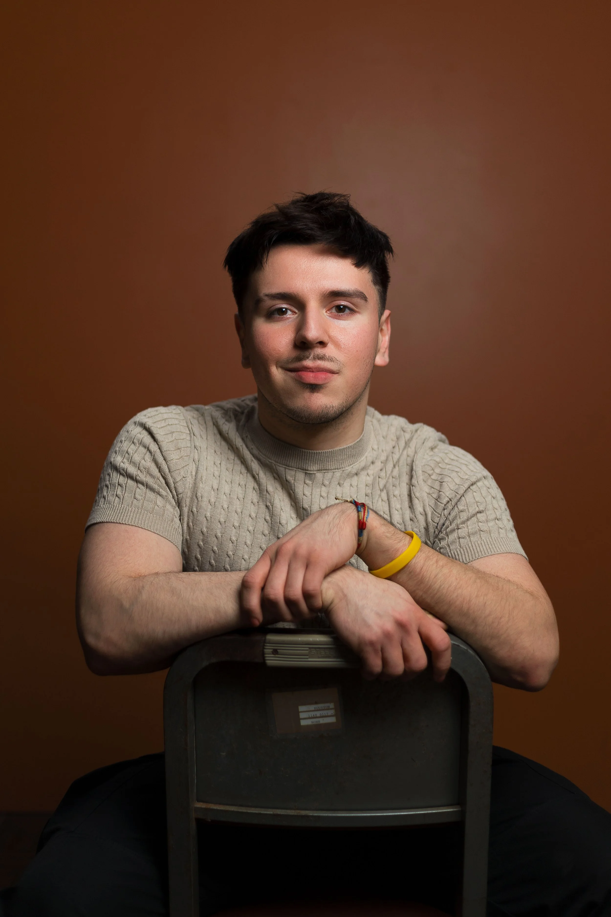 A young man with dark hair and a slight smile, wearing a beige knit sweater, sitting behind a gray chair, with his arms resting on the back of the chair, against a brown background.