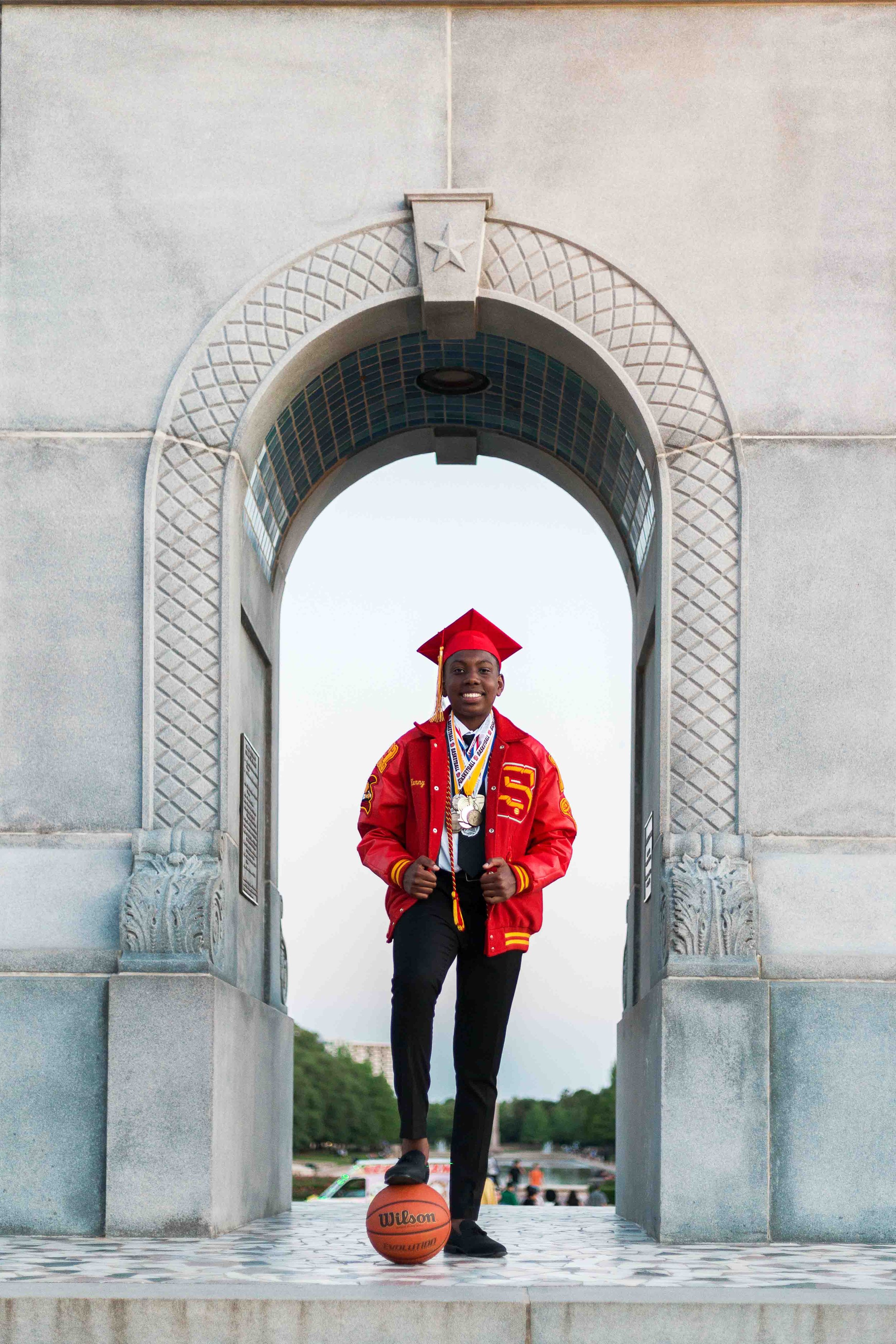 Graduation portrait at Hermann Park in Houston featuring a smiling young man in a red cap and gown with honor medals, standing in a decorative stone archway with a basketball at his feet