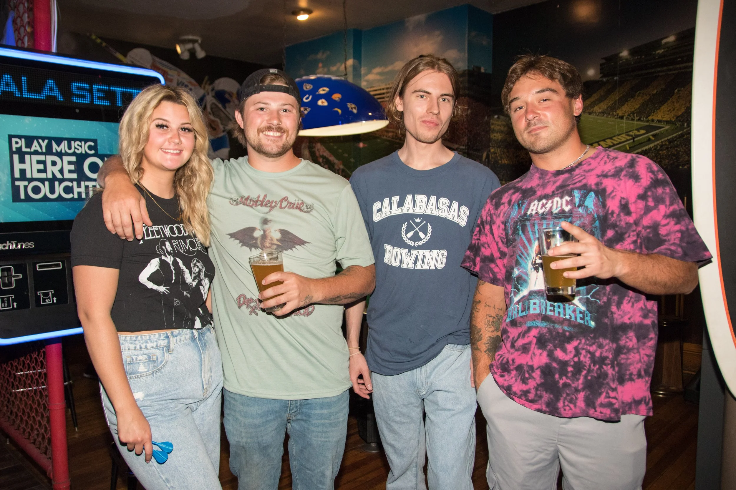 Four young adults standing together in a bar, smiling, with drinks in their hands and casual clothing.