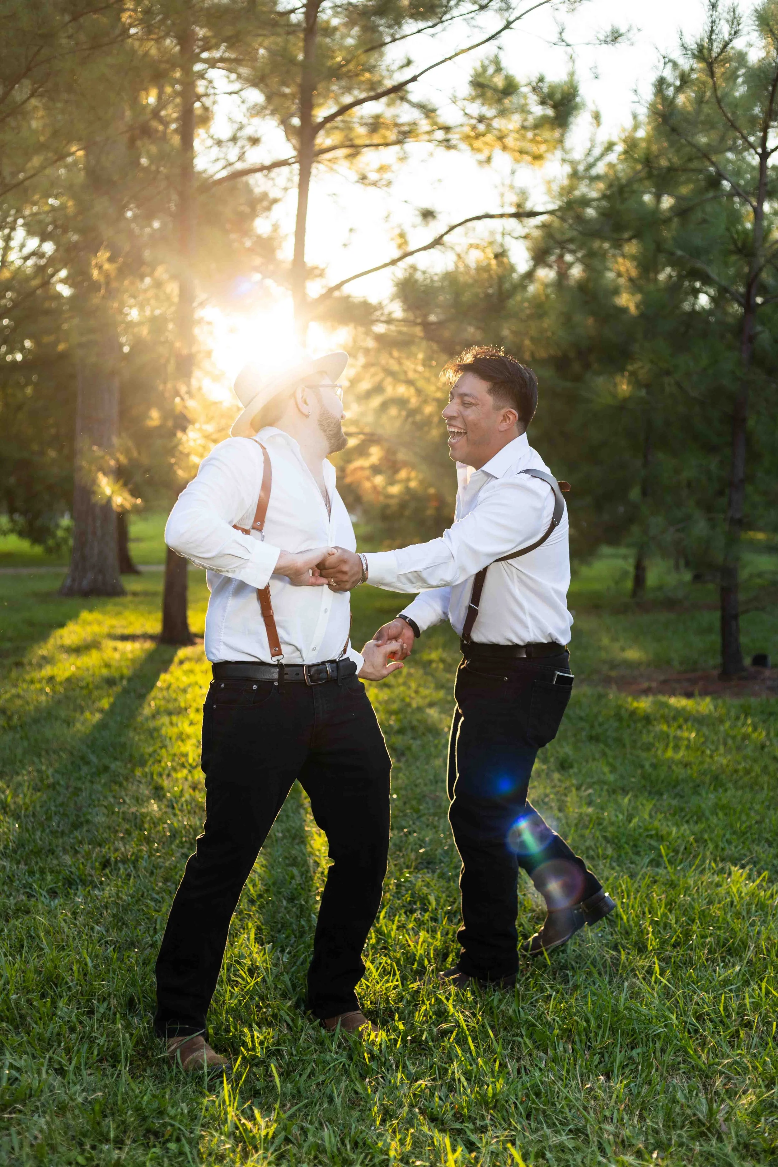 Hermann Park engagement portrait in Houston, Texas by Lev’s Photography featuring two men in white shirts and black pants dancing and laughing outdoors during sunset with trees and green grass in the background.