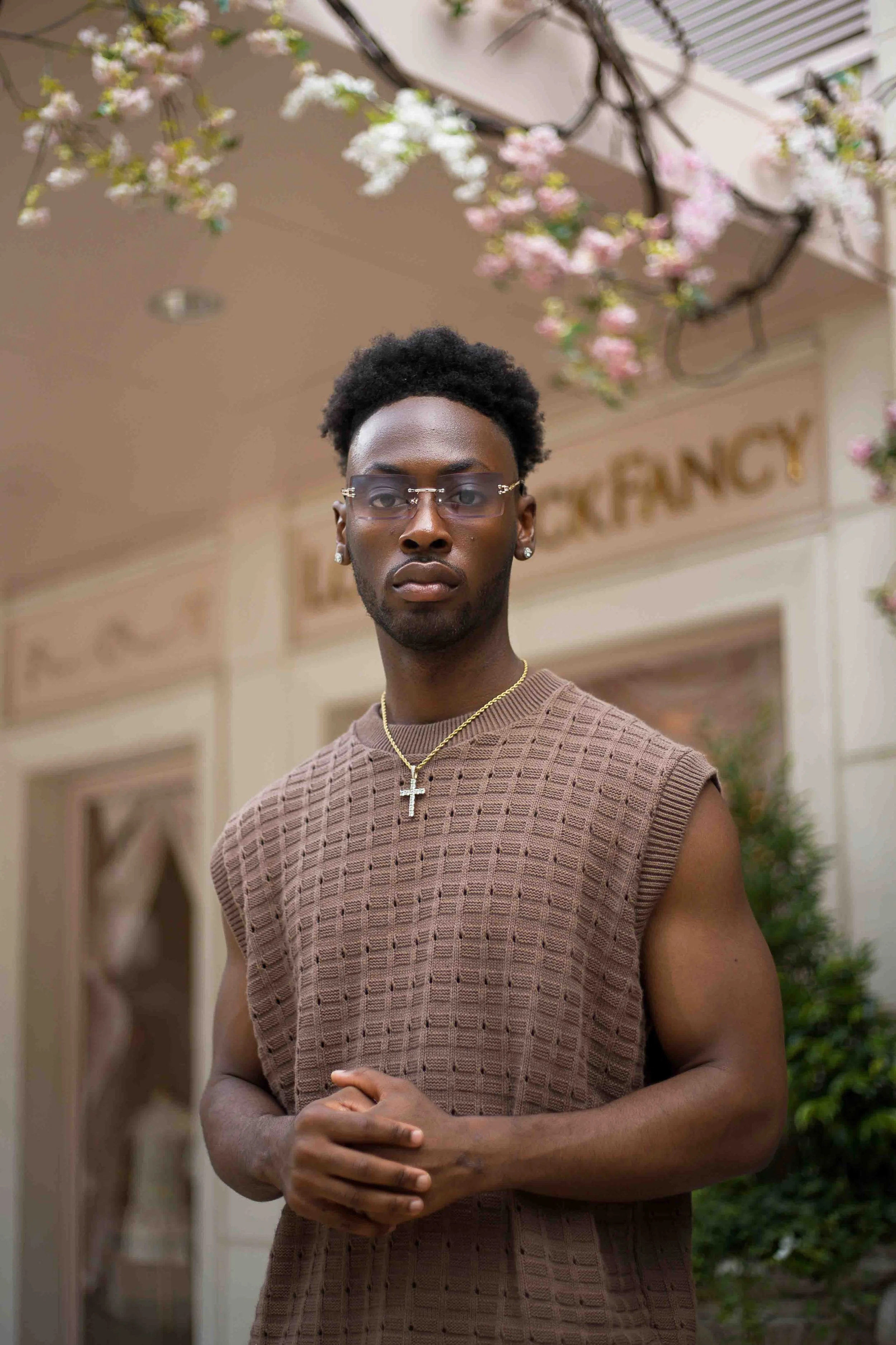 Houston lifestyle fashion portrait by Levs’ Photography featuring a young man with dark skin, wearing glasses, a gold cross chain, and earrings, standing outdoors in front of a building with a ‘LAUNDRY & FANCY’ sign and pink and white flowers overhea