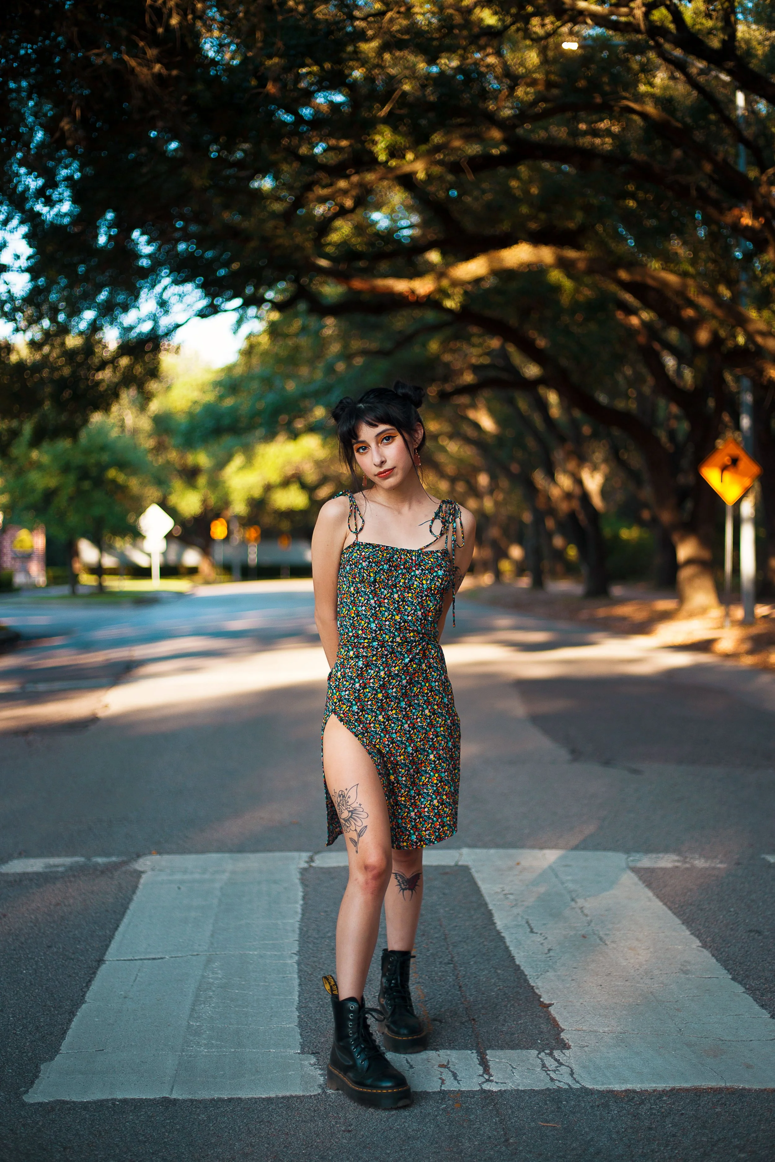 A young woman with dark hair styled in two buns, wearing a colorful floral dress and black combat boots, standing on a crosswalk on a tree-lined street during daylight.