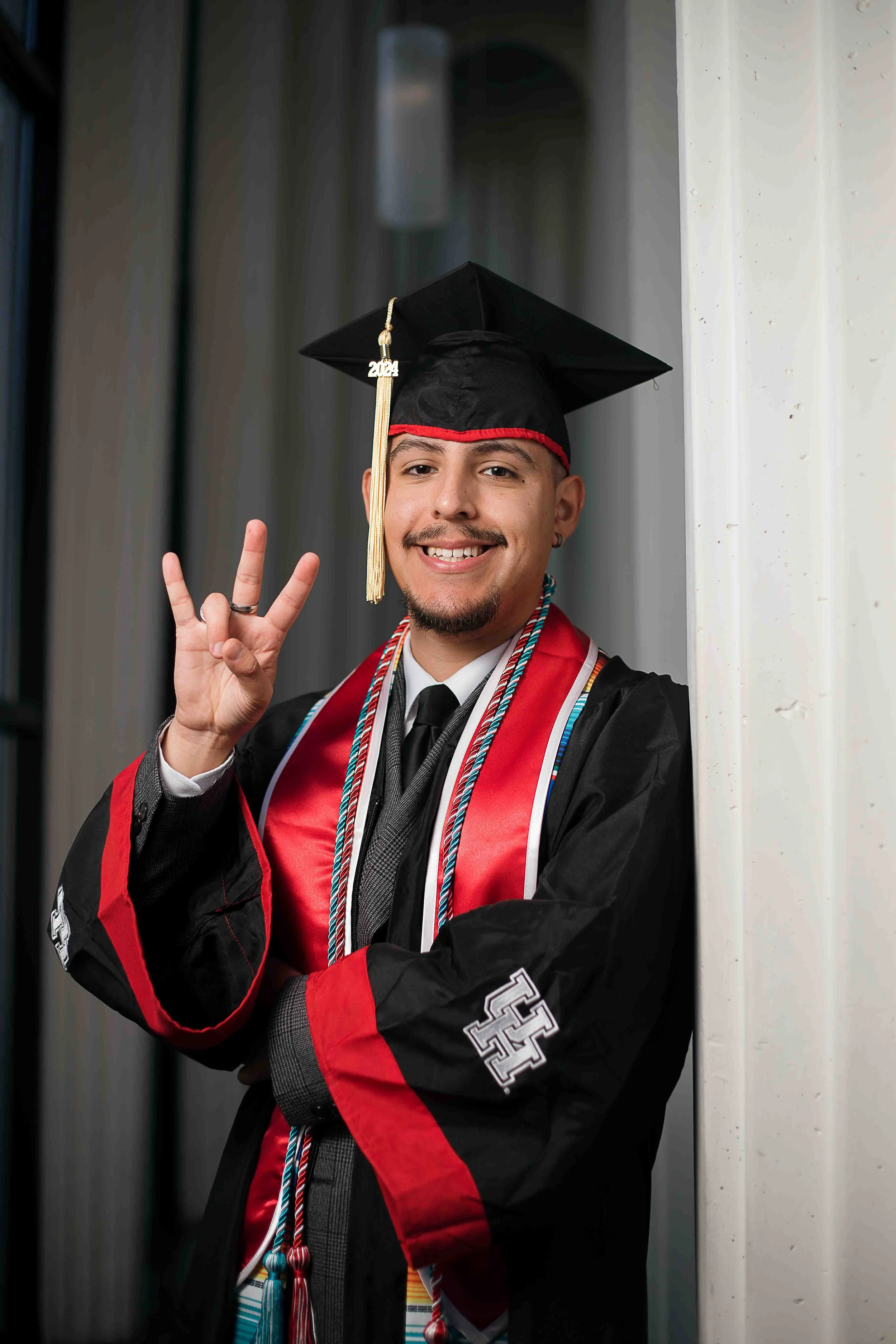 Candid graduation portrait of a University of Houston student wearing a black cap and gown with red accents and honor cords, smiling and flashing a peace sign with his right hand