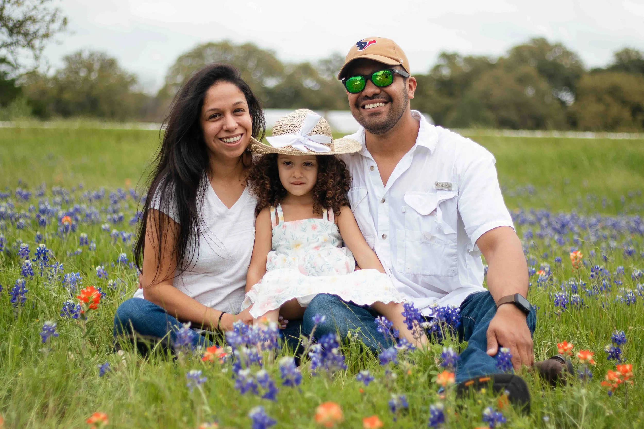 Bluebonnet family portrait in Texas by Lev’s Photography featuring a family of three sitting in a field of wildflowers with trees in the background, smiling and enjoying time outdoors