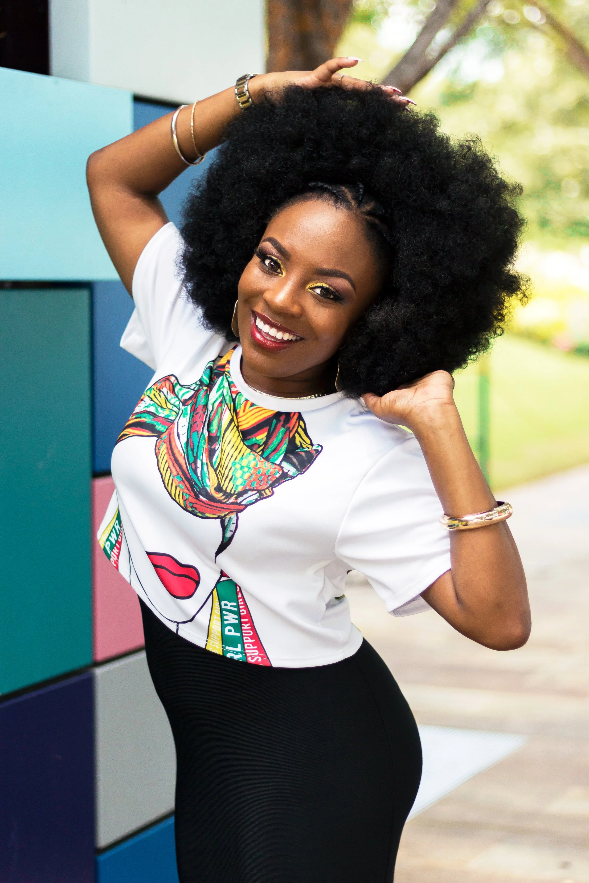 A woman with large curly black hair smiling outdoors, wearing a colorful graphic t-shirt and black pants, standing near multicolored lockers.