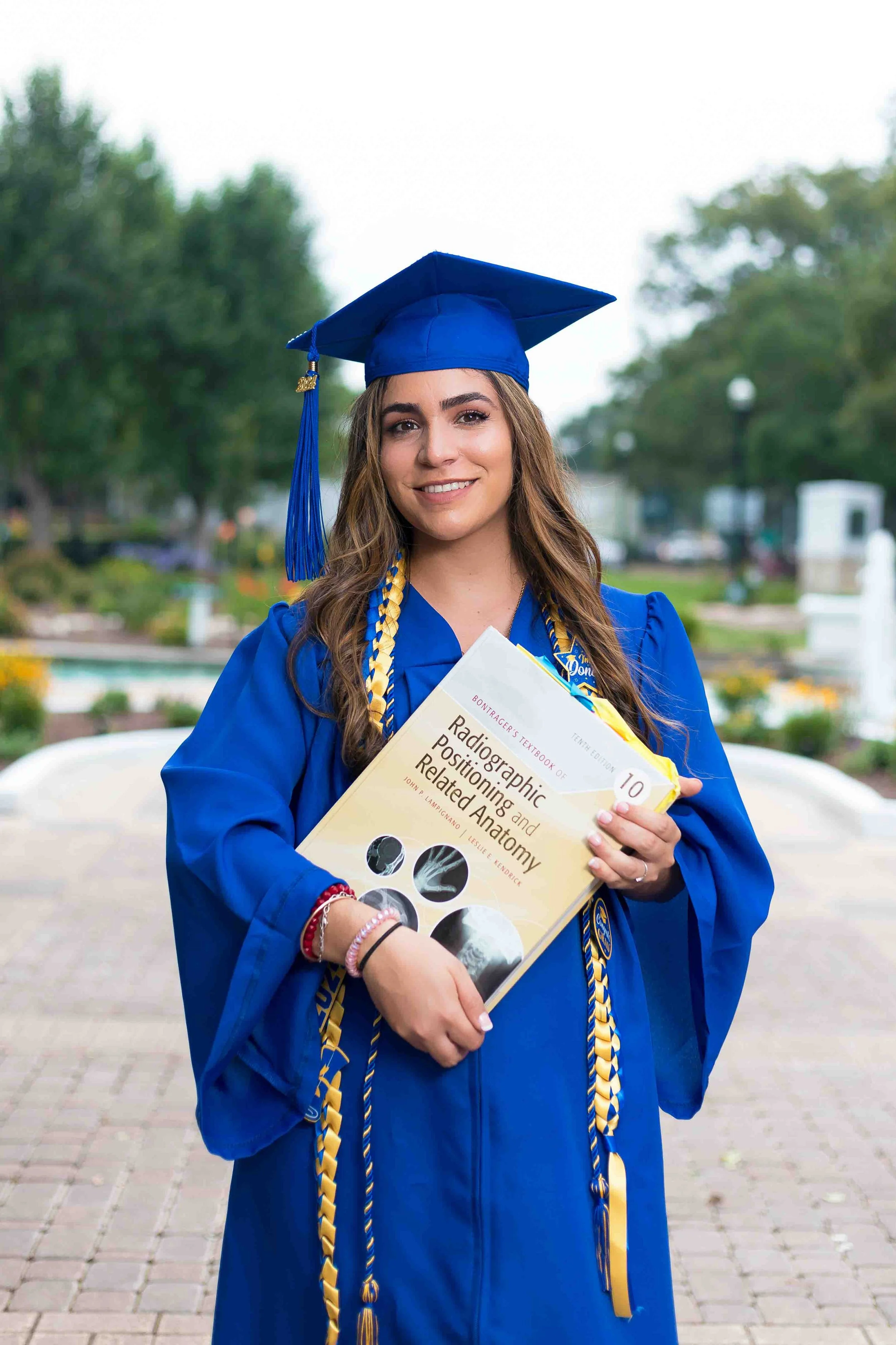 Graduation portrait of a young woman in a blue cap and gown holding a textbook titled Radiographic Positioning and Related Anatomy while standing on a tree-lined campus path at Helen’s Garden