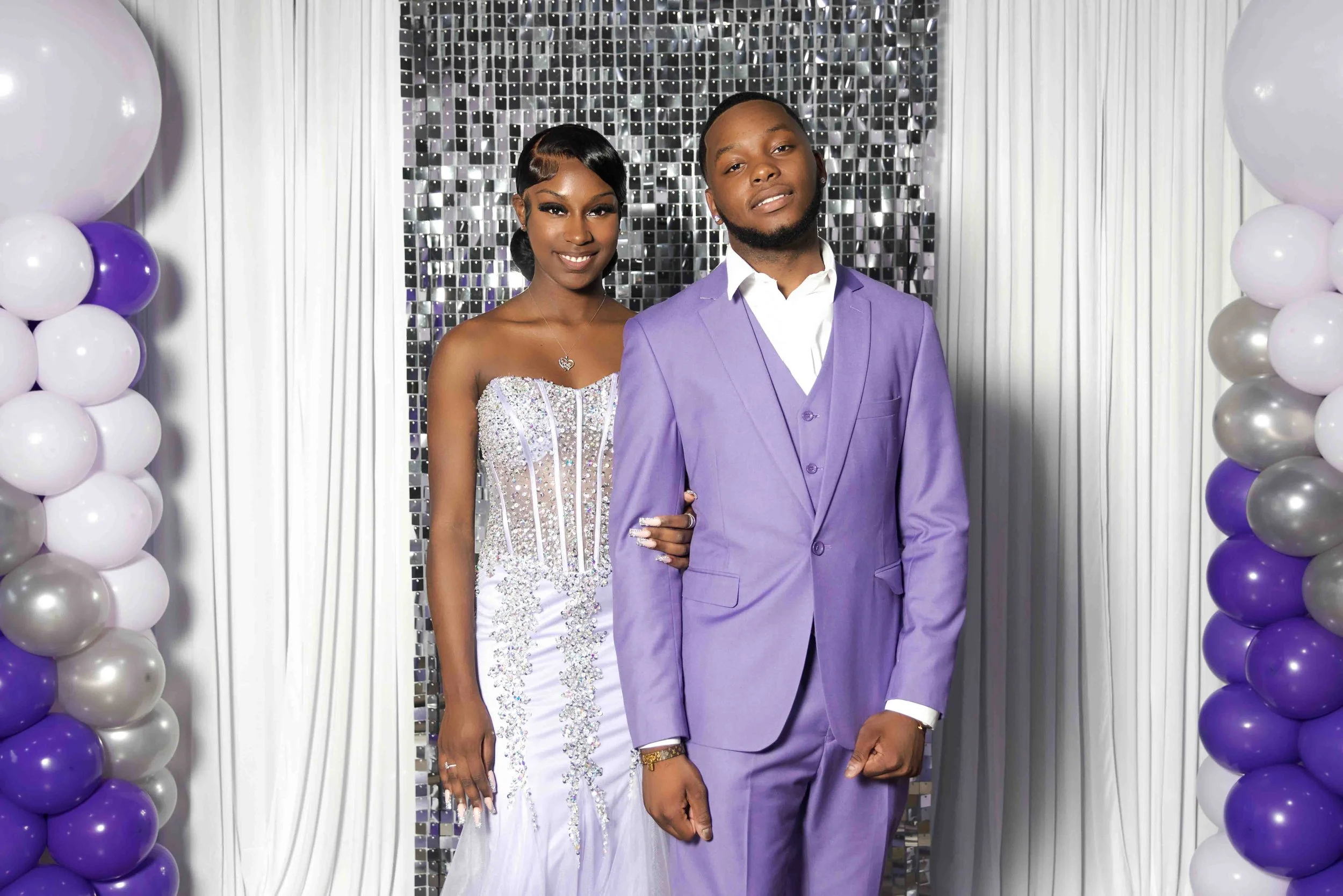 Prom portrait of a couple dressed in formal attire standing in front of a decorated backdrop with balloons and metallic tiles, featuring a woman in a strapless bejeweled white gown and a man in a purple suit