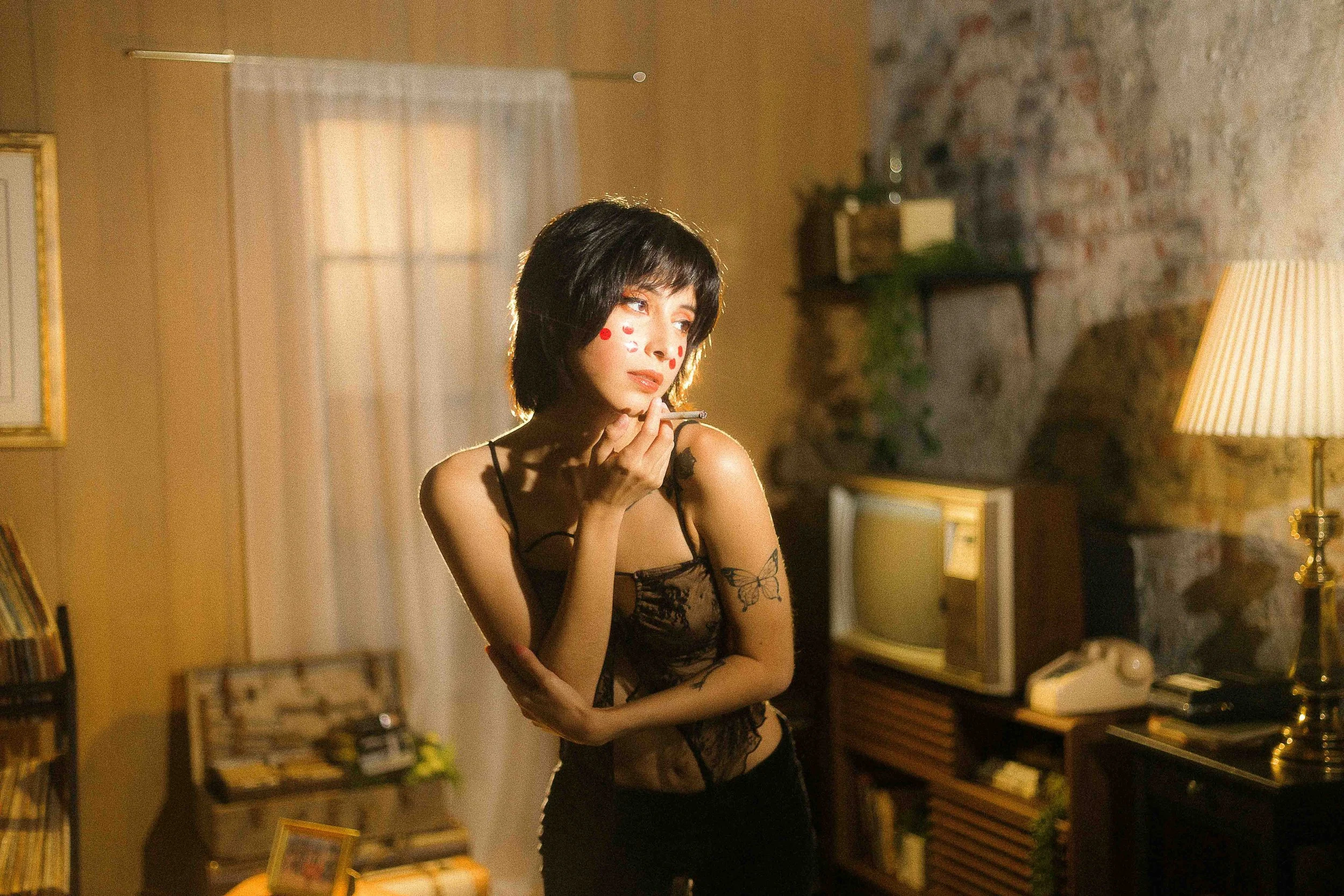 Editorial creative portrait of a woman with short black hair and red dot makeup applying lipstick in a dimly lit vintage-style room with a lamp, television, and shelves in the background