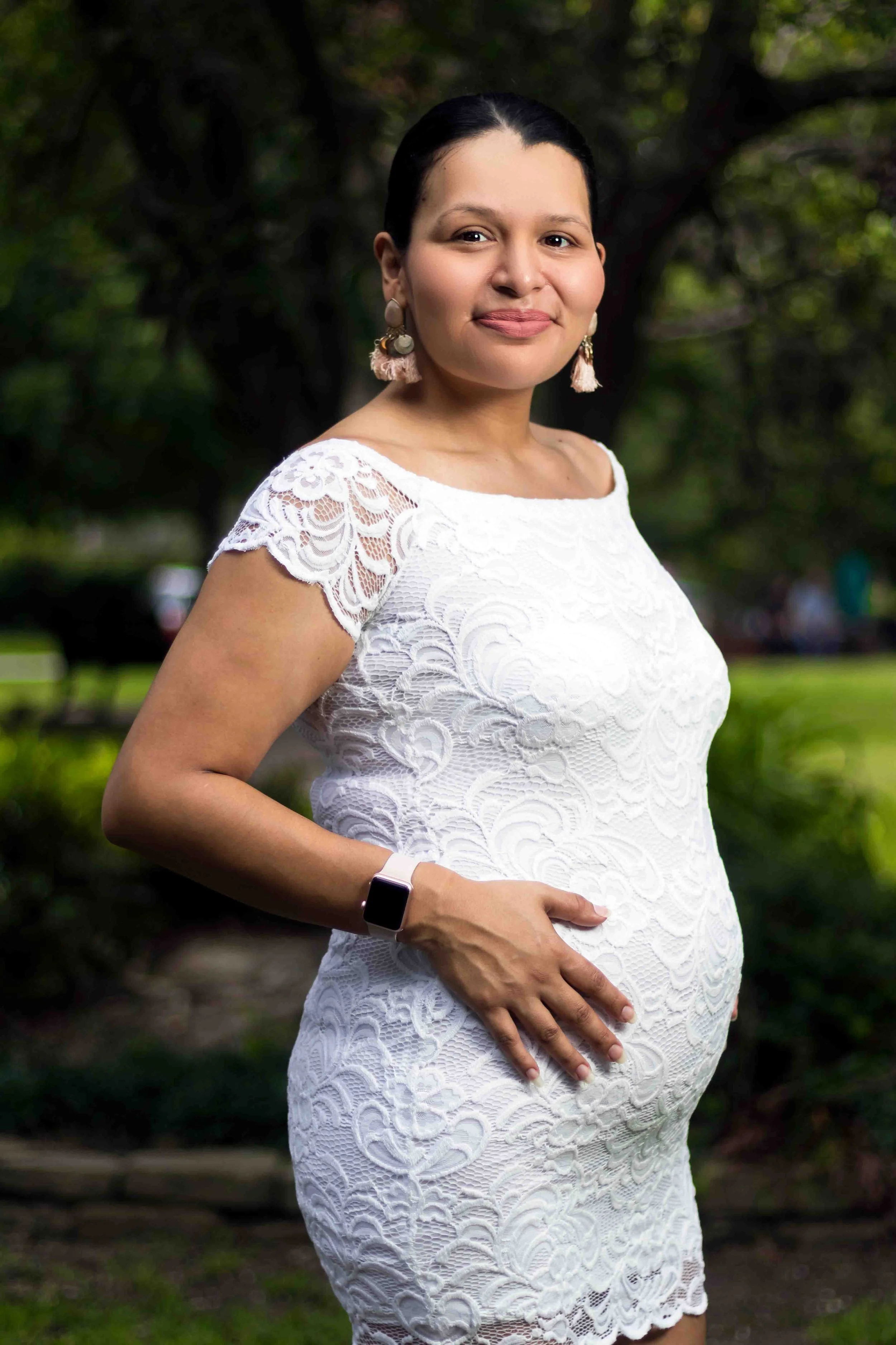 Maternity portrait session in The Heights, Houston, Texas by Lev’s Photography featuring a pregnant woman with dark hair in a white lace dress standing outdoors in a green park.