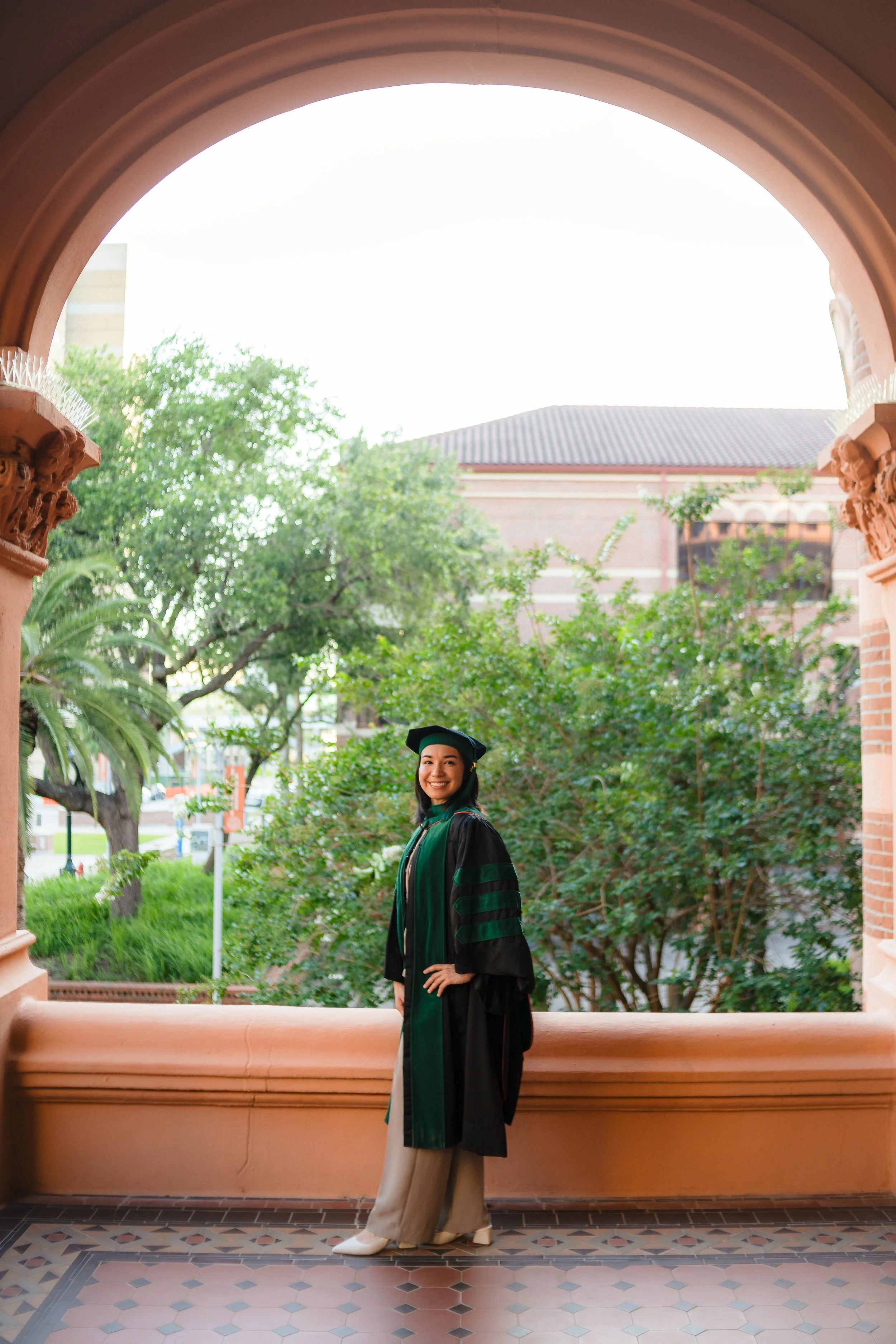 A woman in a graduation gown and cap standing in an arched architectural setting with a view of trees and buildings in the background.
