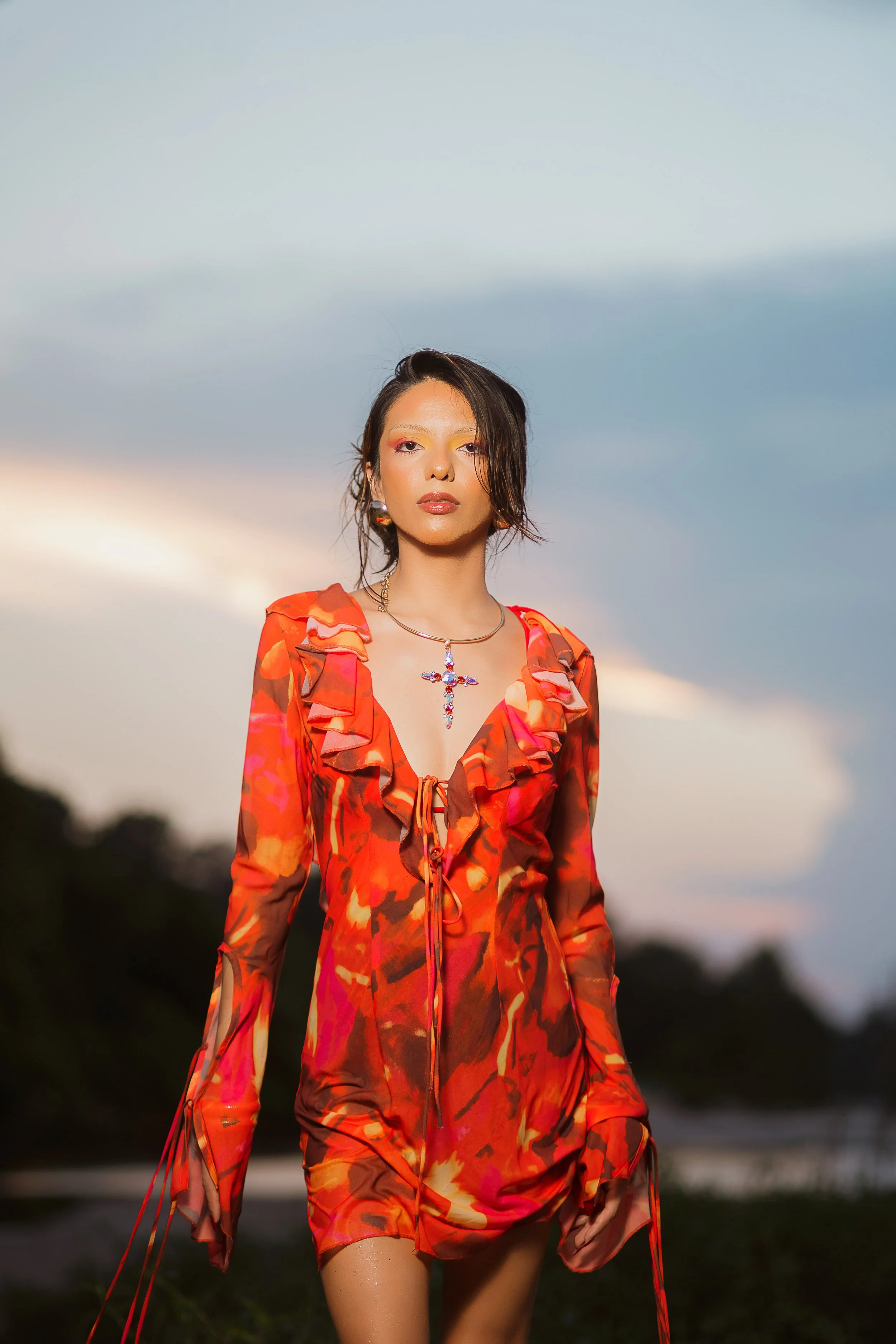 A woman with dark, wavy short hair wearing a colorful, ruffled red, orange, and pink patterned dress, standing outdoors during sunset with a sky and some trees in the background.