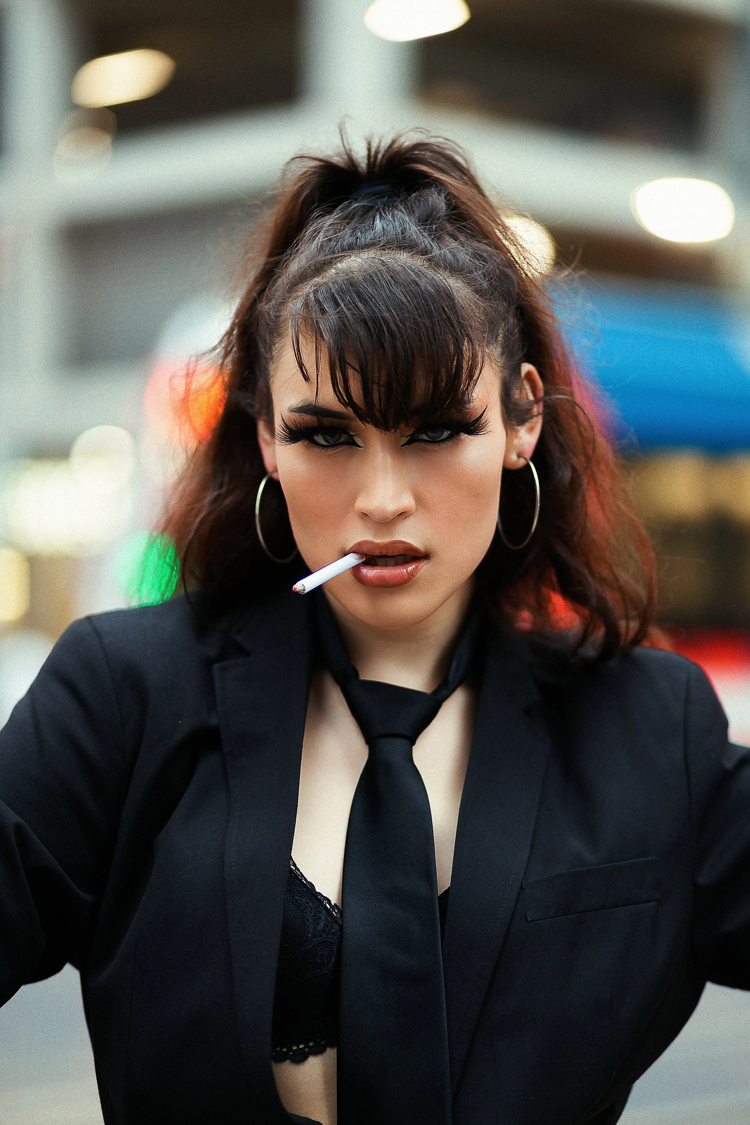 A woman with dark hair, wearing hoop earrings, a black blazer, and a loosened black tie, has a cigarette in her mouth and is looking directly at the camera with a serious expression. The background is blurred with colorful lights.
