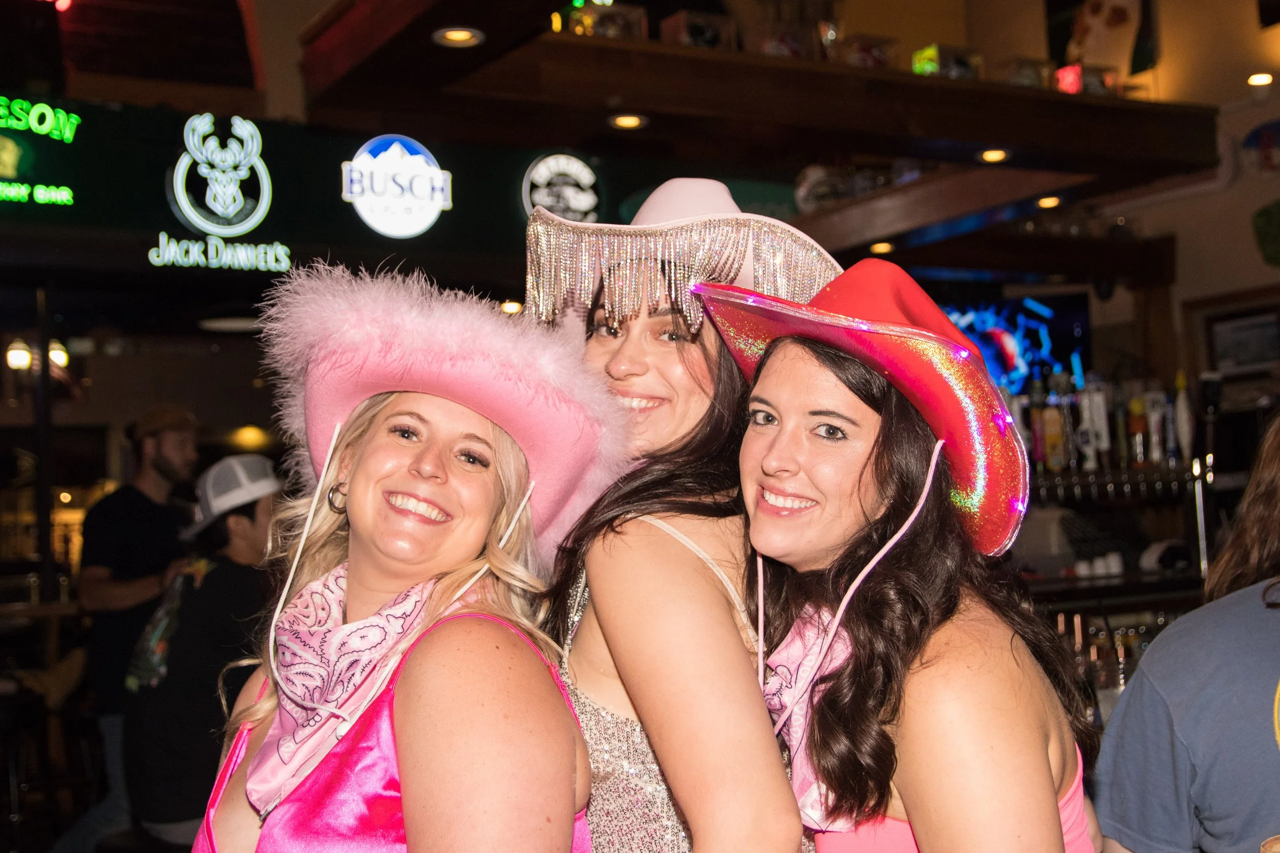 Three women wearing colorful cowboy hats and pink outfits at a bar or party, smiling for the camera.