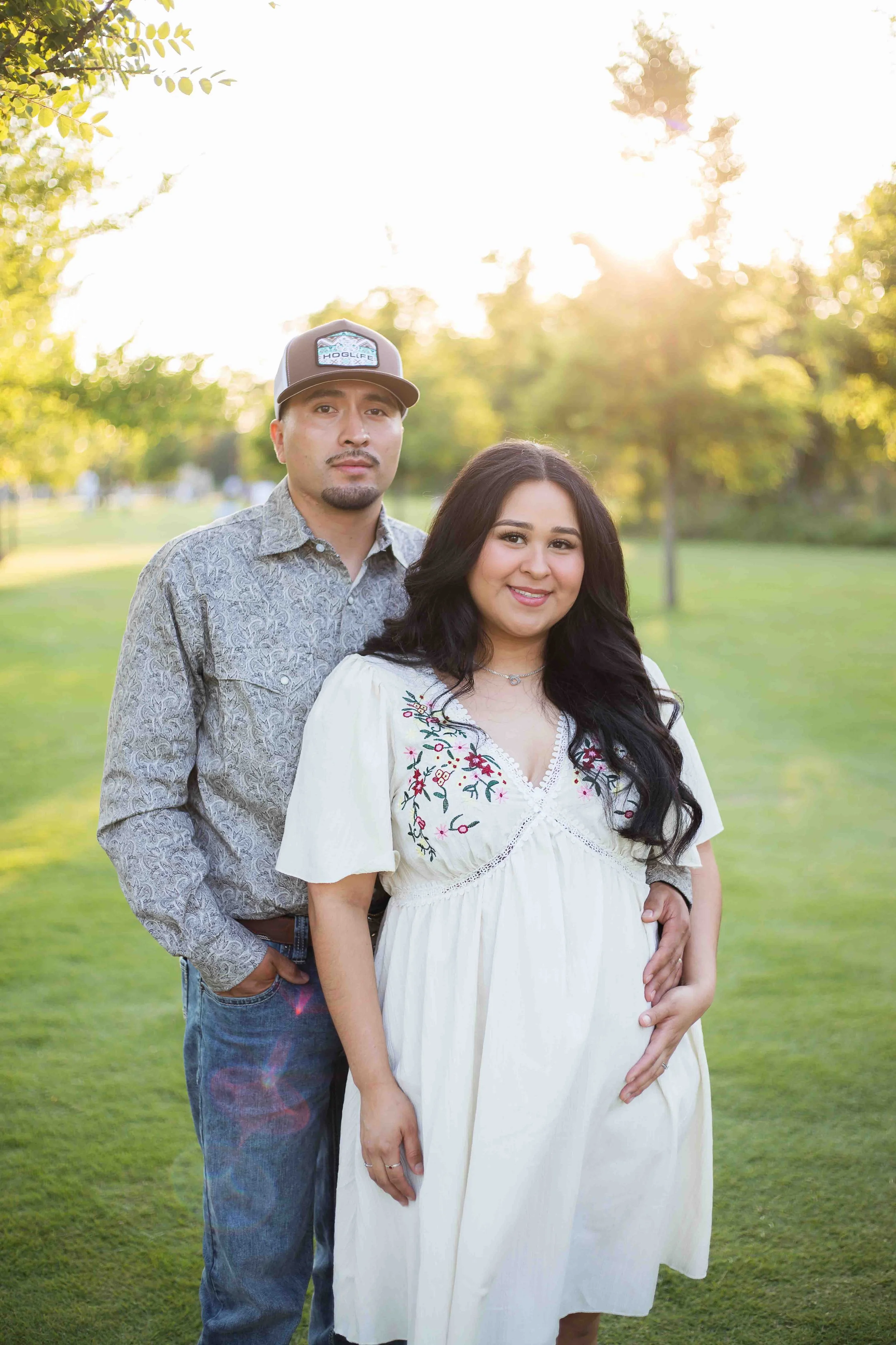 A couple posing outdoors in a park during sunset. The woman is pregnant and wearing a white dress with floral embroidery. The man is dressed in a patterned shirt, blue jeans, and a cap. They are standing on green grass with trees and sunlight in the