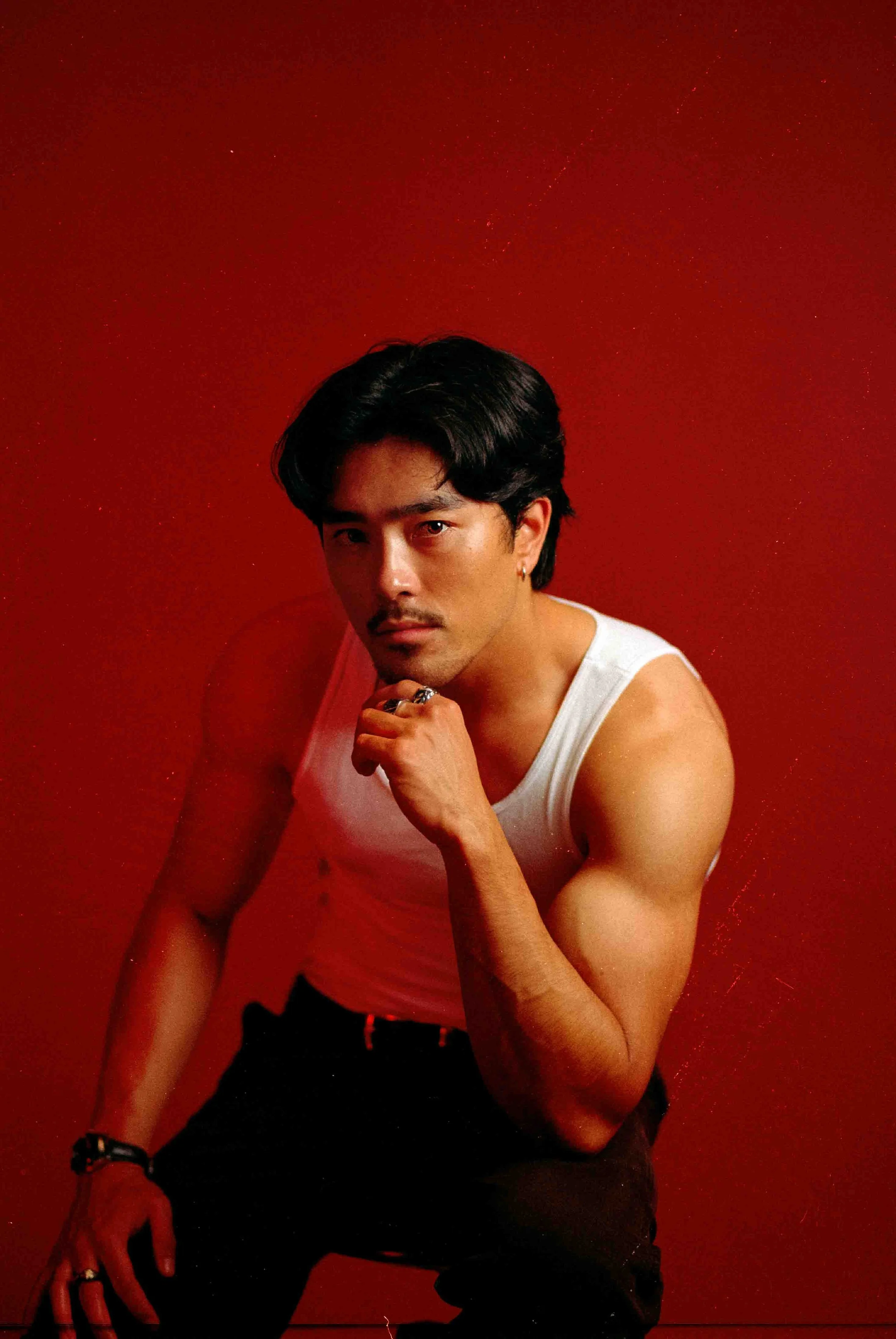 35mm film portrait of a man with black hair wearing a white sleeveless shirt and black pants, sitting against a bold red background with a contemplative expression, resting his chin on his hand
