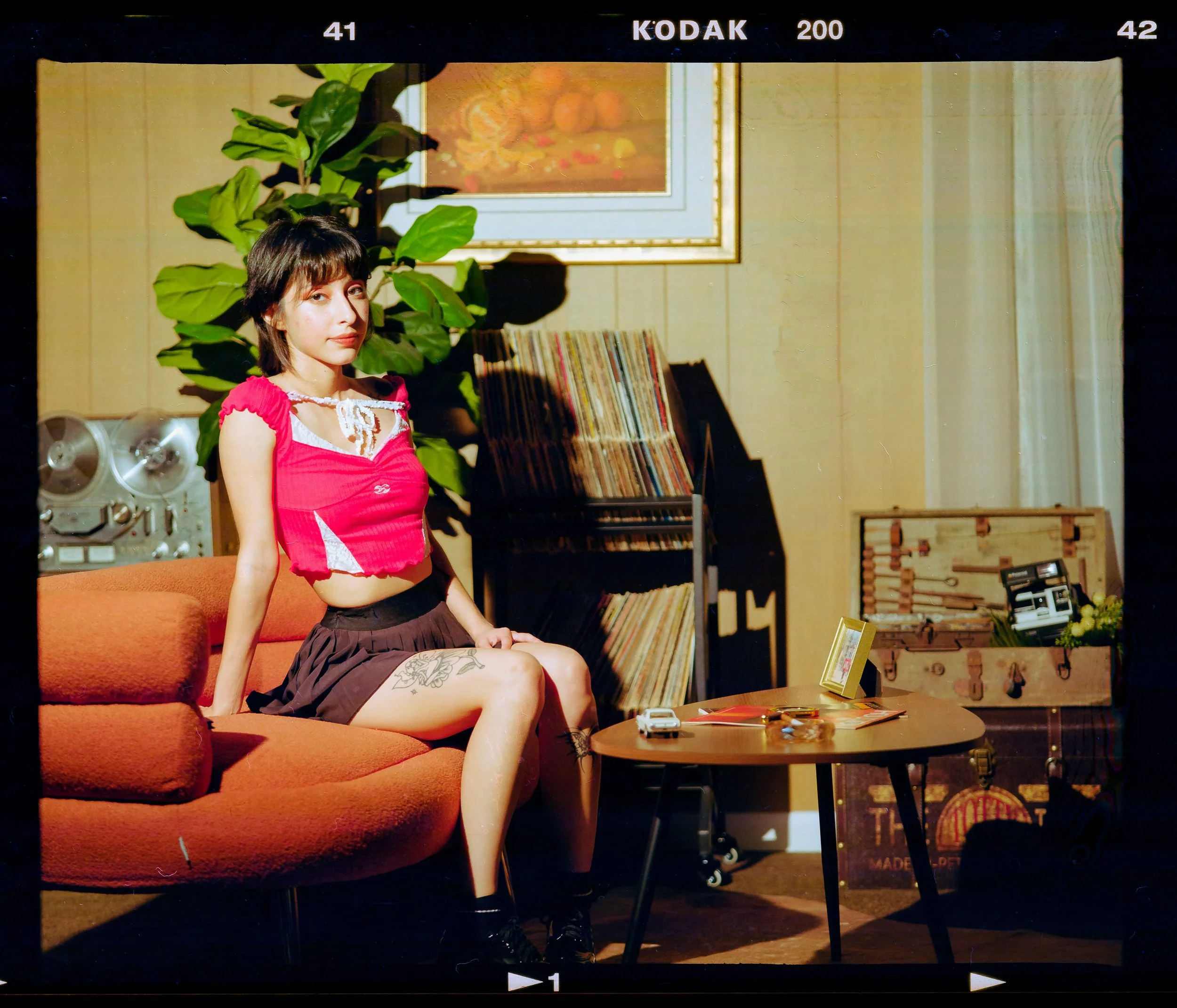 A young woman with short dark hair and tattoos on her left thigh and right leg, sitting on an orange couch in a cozy room with vintage decor. She is wearing a red crop top with white accents and a brown skirt, looking towards the camera. Behind her a