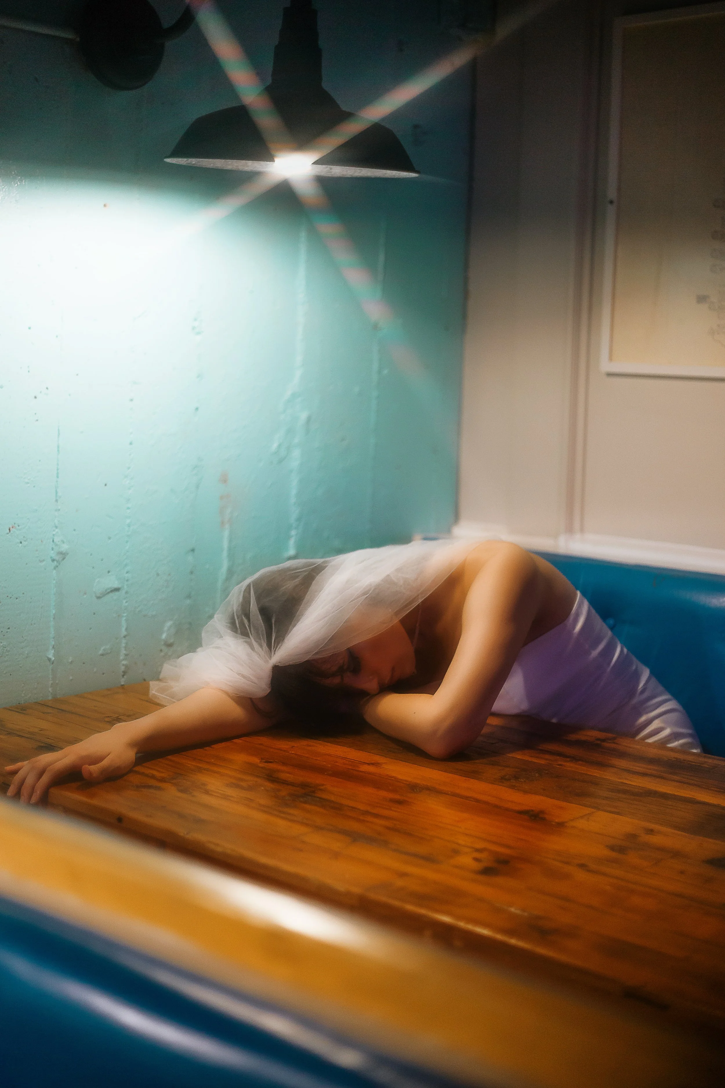 A woman wearing a white dress and veil, possibly a bride, is resting her head and arm on a wooden table in a dimly lit room with a greenish wall and a black hanging lamp.