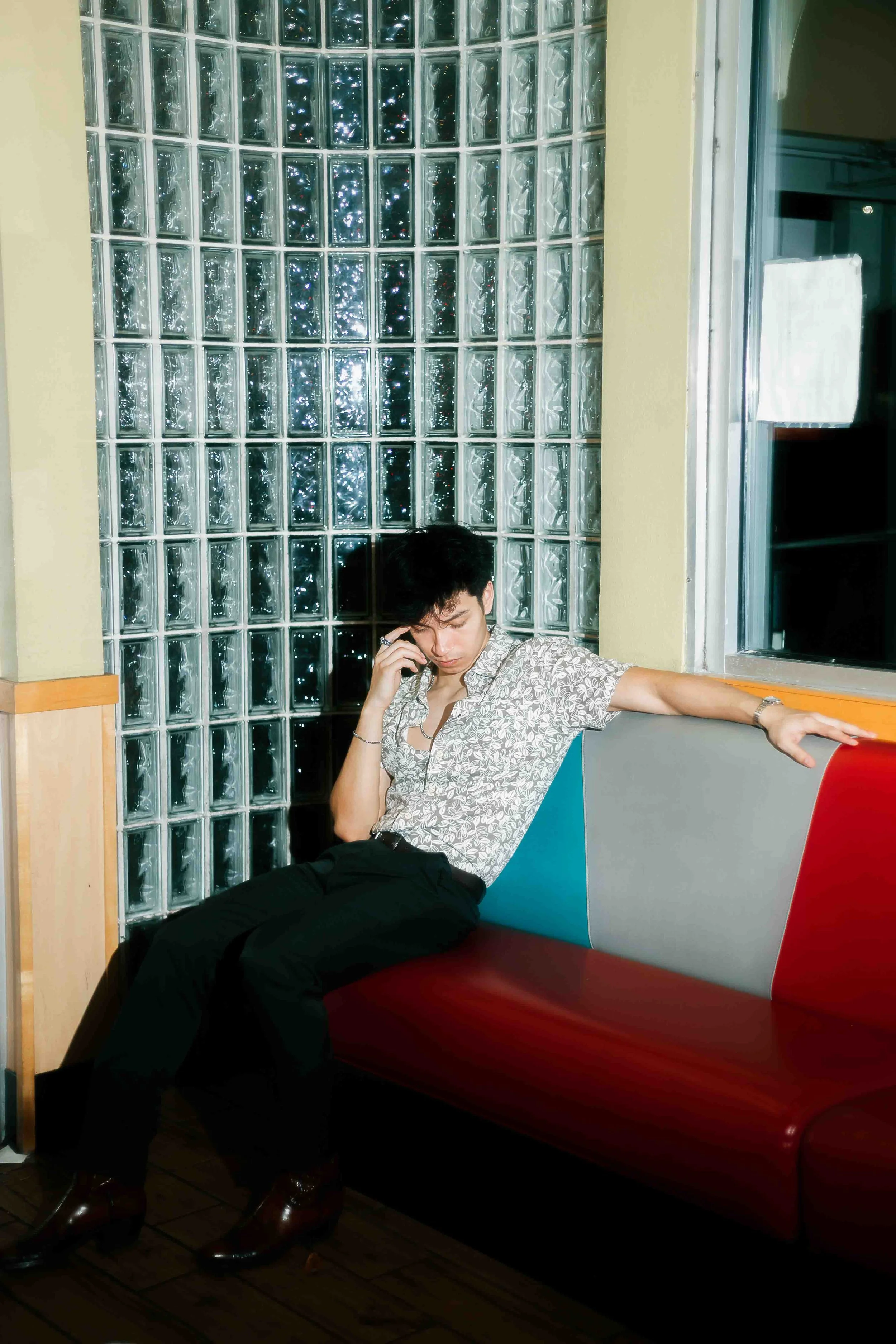 Houston editorial portrait of a young man with black hair wearing a patterned shirt, black pants, and brown boots, sitting on a bench in a restaurant with a glass block wall and window behind him, looking down while on the phone