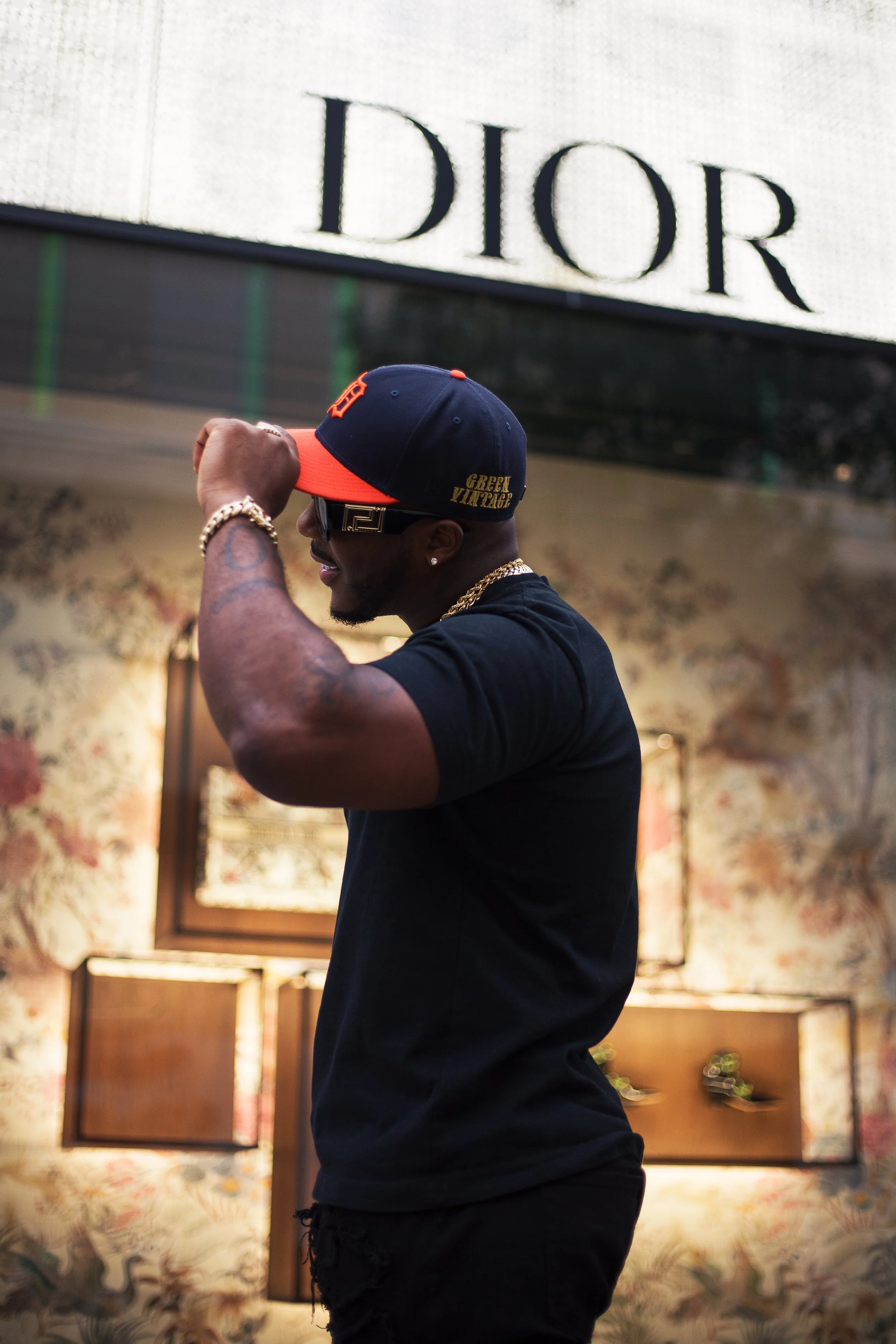 A man wearing a black t-shirt and a navy and orange cap, adjusting the cap, standing in front of an interior wall with floral wallpaper and framed artwork, with a large illuminated Dior sign overhead.