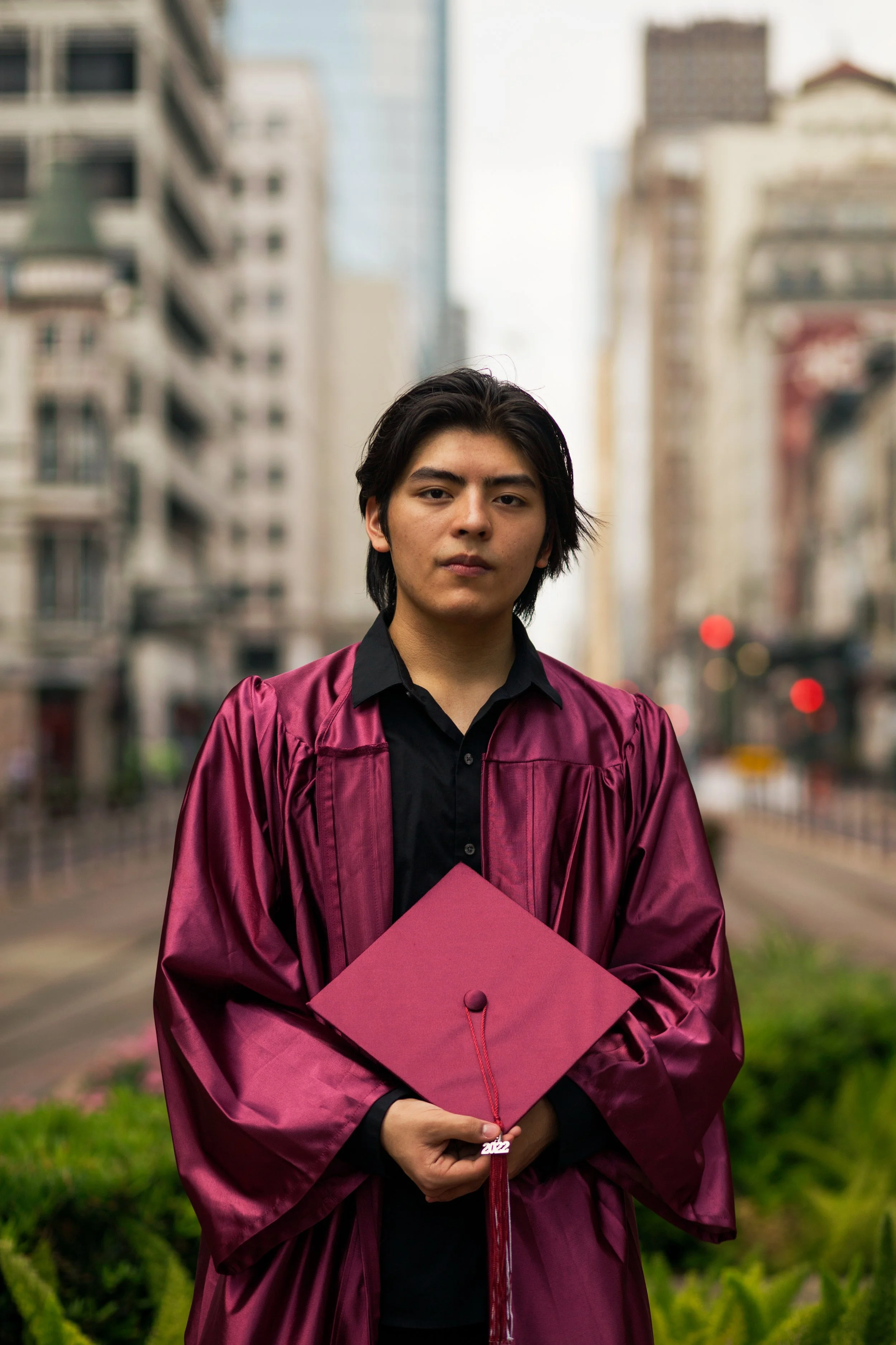 A young man in a maroon graduation gown and cap holding his diploma standing on a city street with tall buildings in the background.