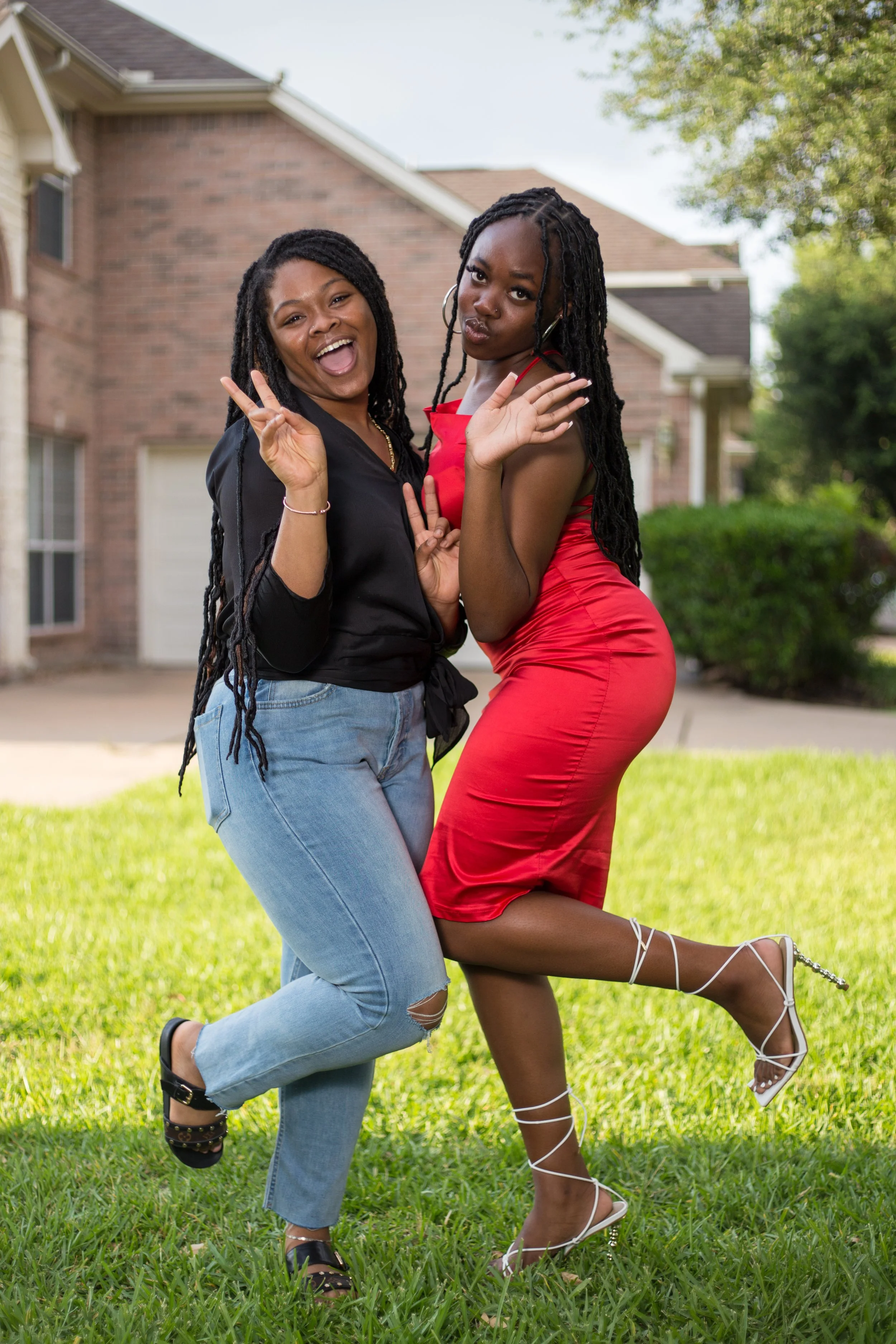 Two women standing on grass, smiling and posing playfully for the camera, with a brick house and trees in the background.