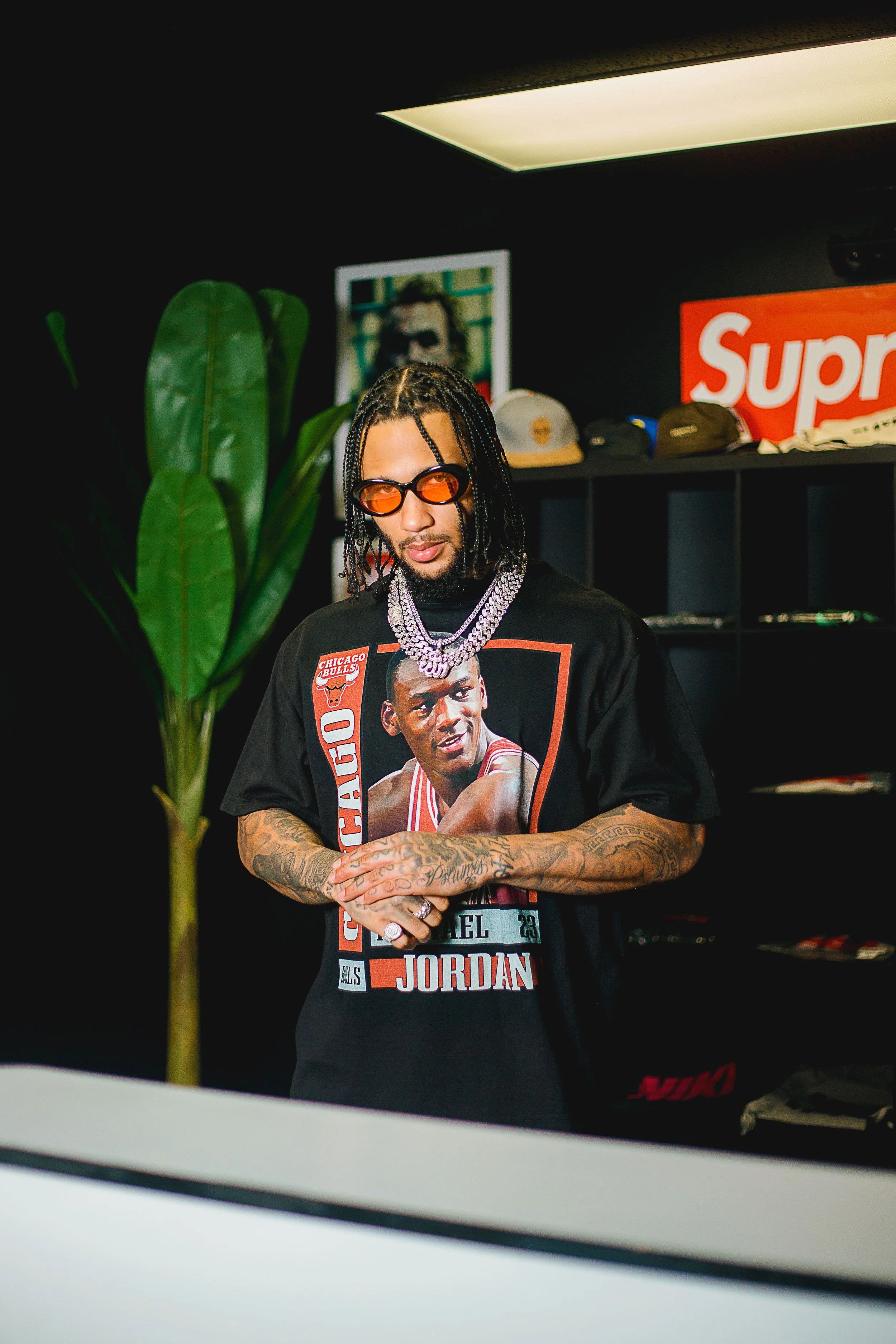 A man with braided hair and tattoos wearing a Chicago Bulls Michael Jordan graphic T-shirt, orange-tinted sunglasses, and layered necklaces, standing indoors in a room with black walls, a large green plant, a framed photo, and shelves with hats, with