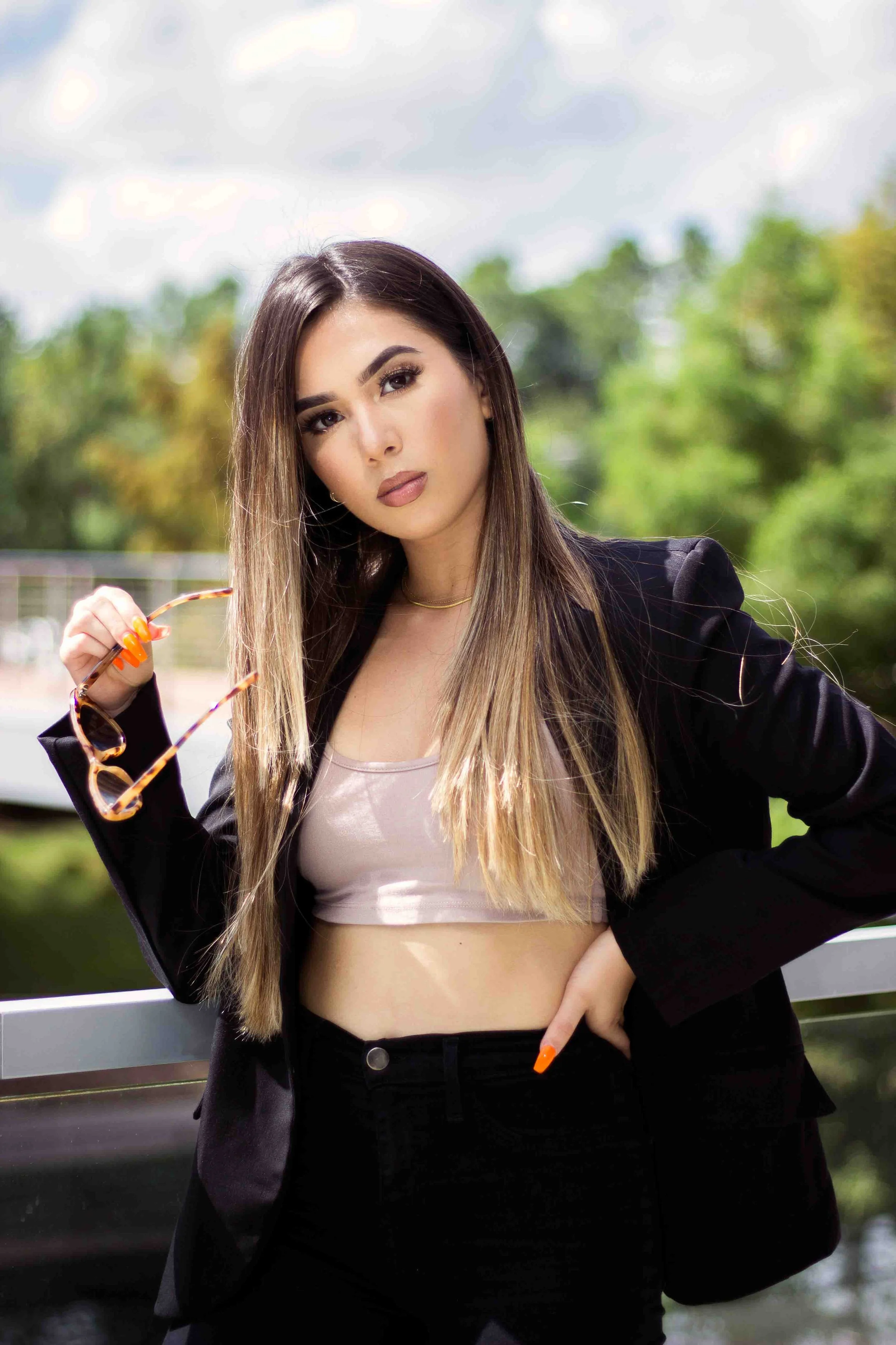 Business branding portrait by Lev's Photography in The Woodlands, Texas featuring a confident woman with long brown hair posing on a bridge, wearing a blazer over a crop top and holding sunglasses, captured outdoors with trees and a dramatic cloudy s