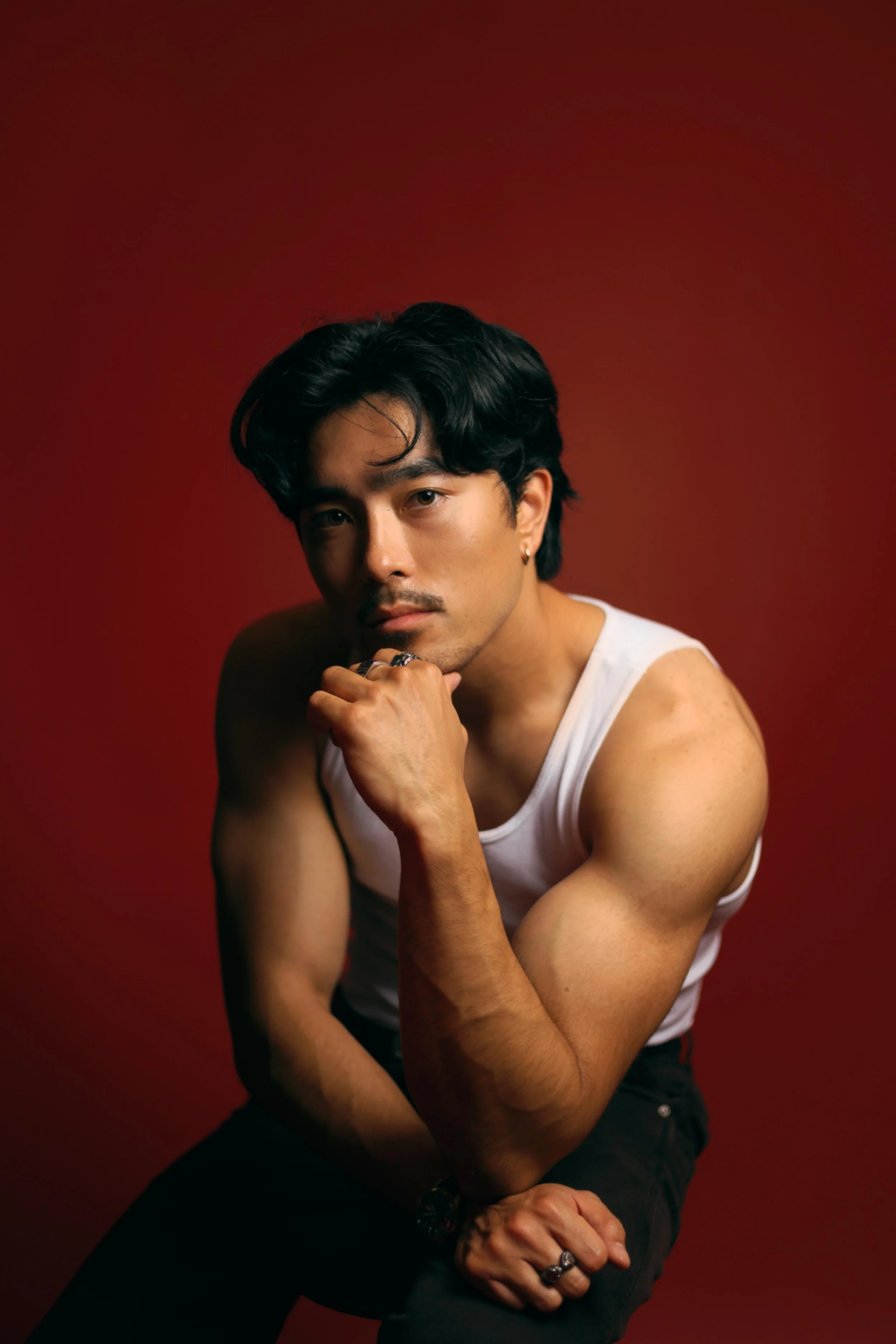 Fitness progress portrait by Lev's Photography in Houston, Texas featuring a man wearing a white tank top, seated against a solid red studio background, showing his physique and progress with professional editorial lighting for a clean and motivation