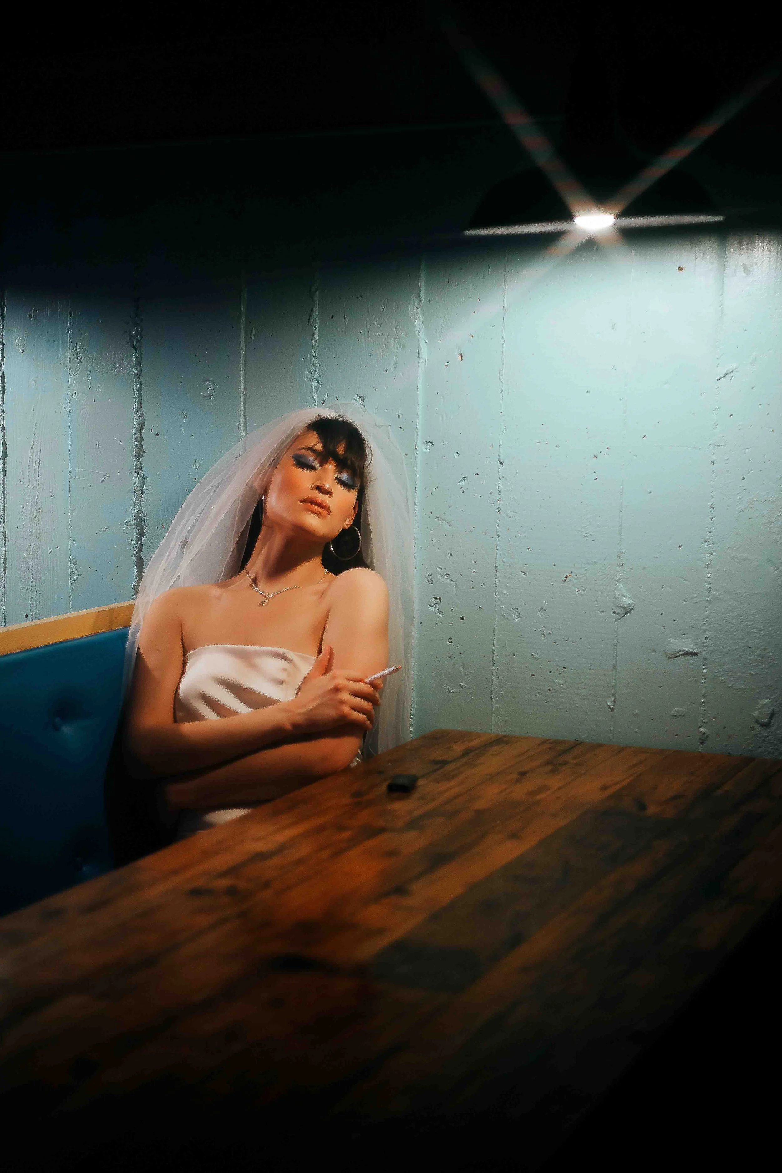 Moody bridal portrait of a woman wearing a veil and wedding makeup, seated at a wooden table holding a cigarette in a dimly lit room with a textured teal wall backdrop