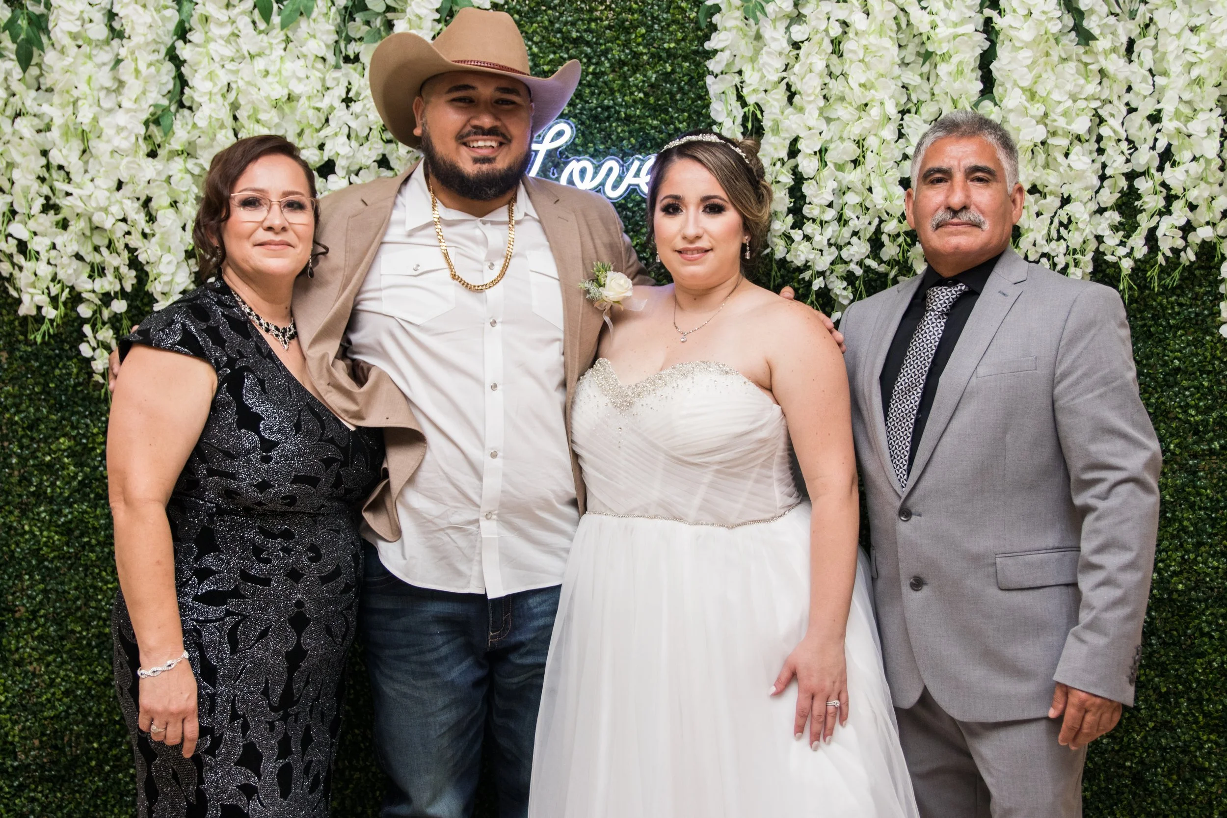 Group of five people at a wedding, standing in front of a green floral backdrop. The bride is in a white wedding dress, and the groom is next to her wearing a beige suit and cowboy hat. Two older people, a woman in a black dress and a man in a gray s