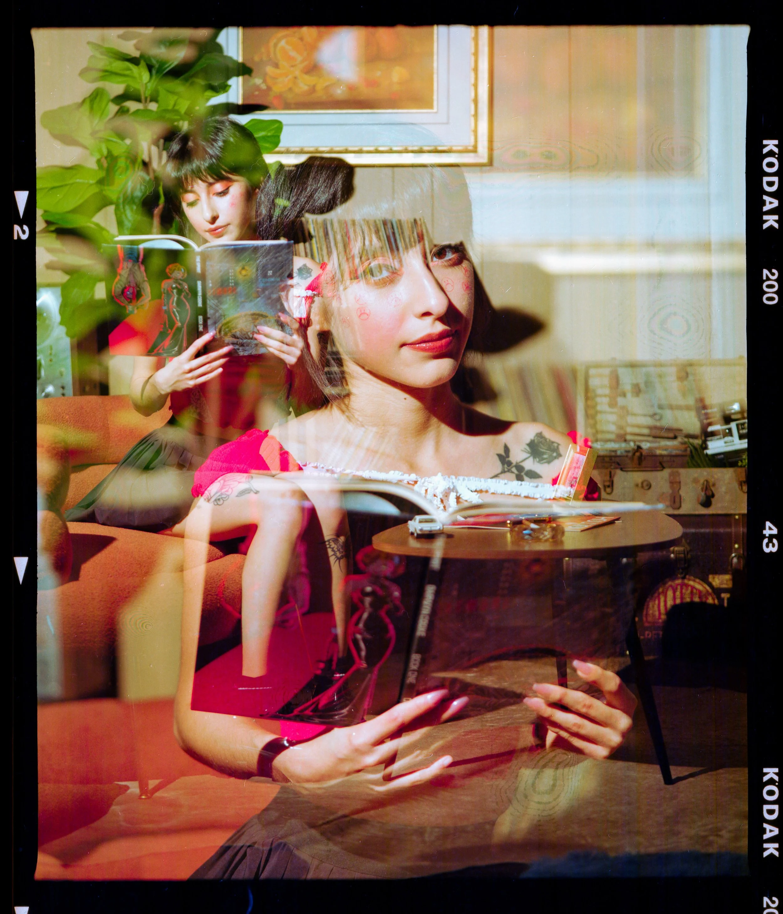 A woman with dark brown hair and a tattoo on her shoulder, holding a magazine, sitting on a sofa. A reflection shows another woman with short dark hair reading a book, with a plant and framed artwork in the background.