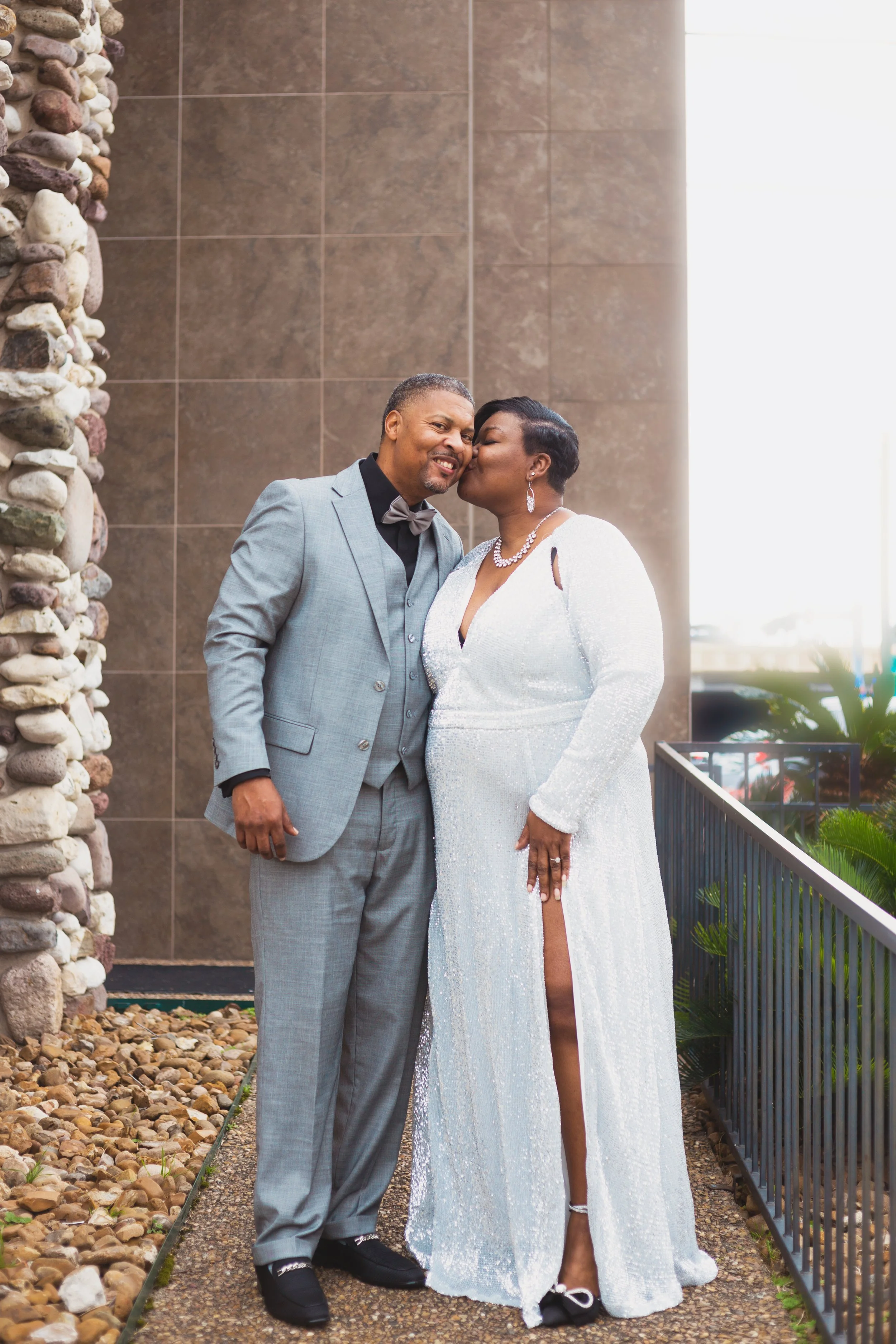 A couple dressed in formal attire, the woman in a white sequin gown and the man in a light grey suit, sharing a kiss on the cheek outdoors near a stone wall and railing.