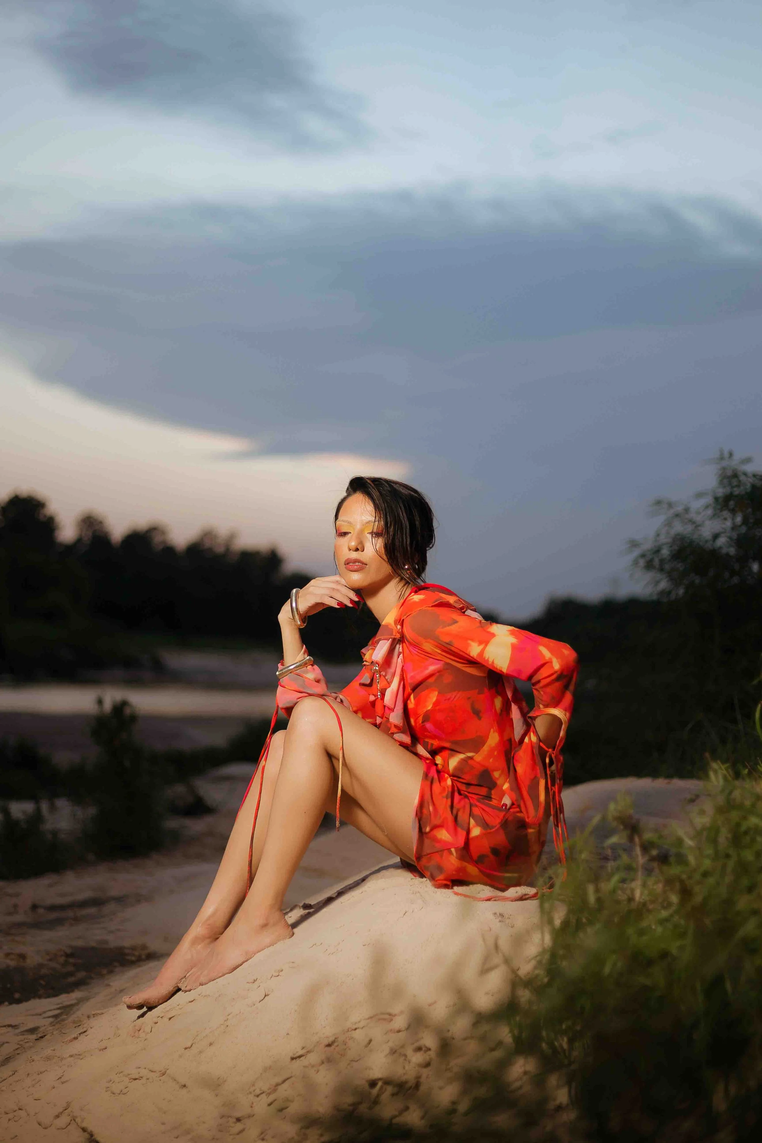 Outdoor portrait of a woman in a red and orange patterned dress sitting on the sand at dusk, with trees and a cloudy evening sky in the background in Humble